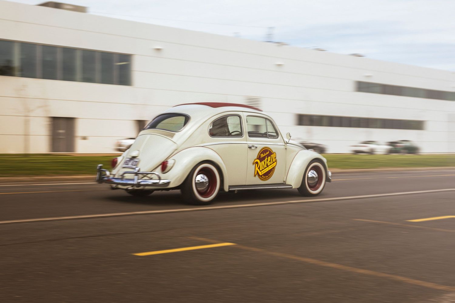 Cream vintage Volkswagen Beetle with whitewall tires and a round logo on the door driving past a large industrial building.