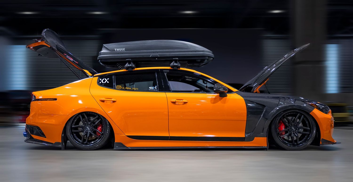 A custom orange sport sedan with carbon fiber panels and a rooftop cargo box has its hood and trunk raised at a car show.