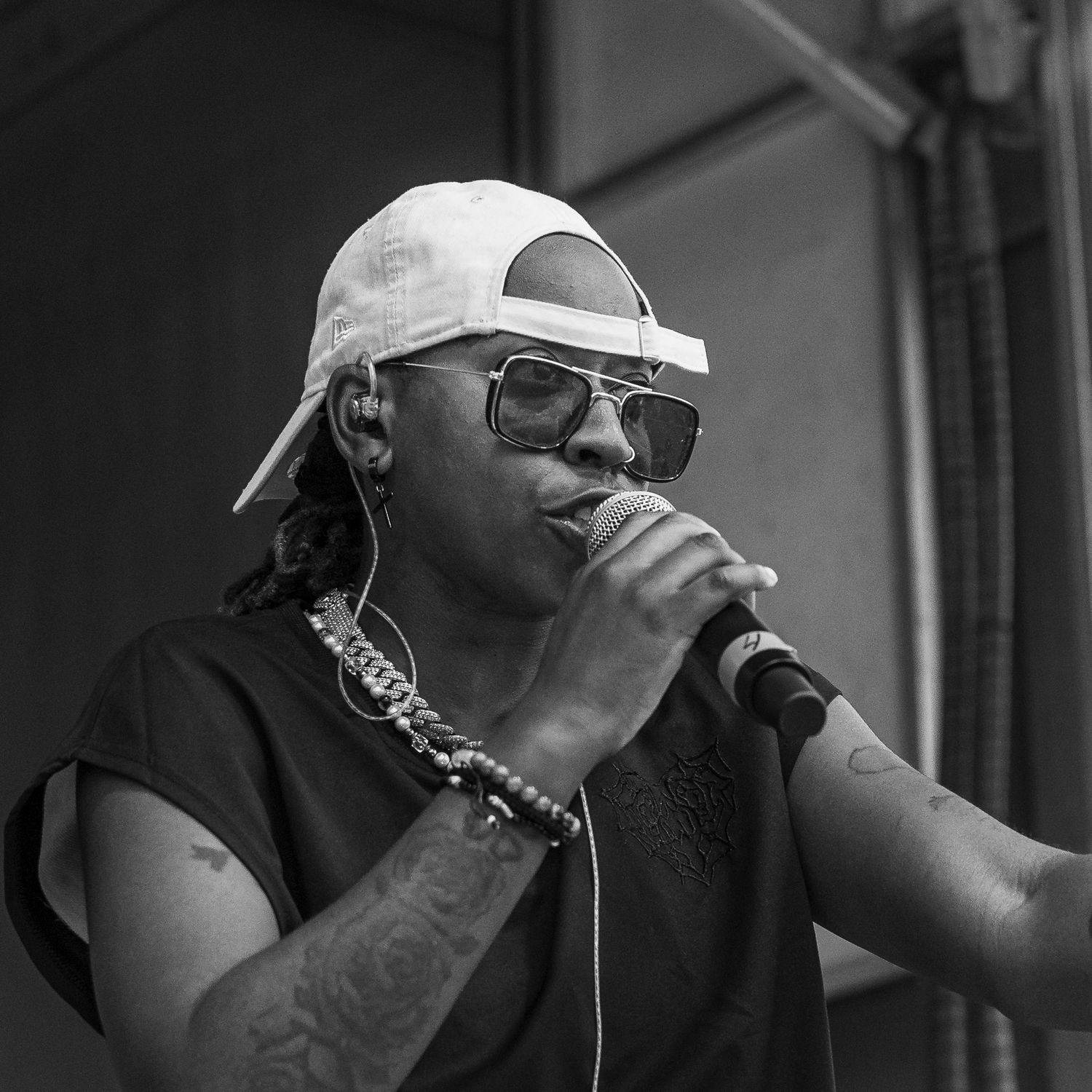 Black and white photo of a performer in a white cap and sunglasses singing into a microphone with tattooed arms.