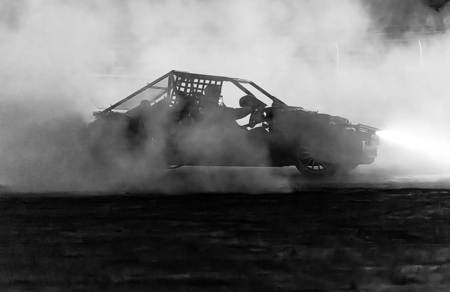 Monochrome shot of a race car with a roll cage spinning its tires in heavy smoke with headlights glowing at night.
