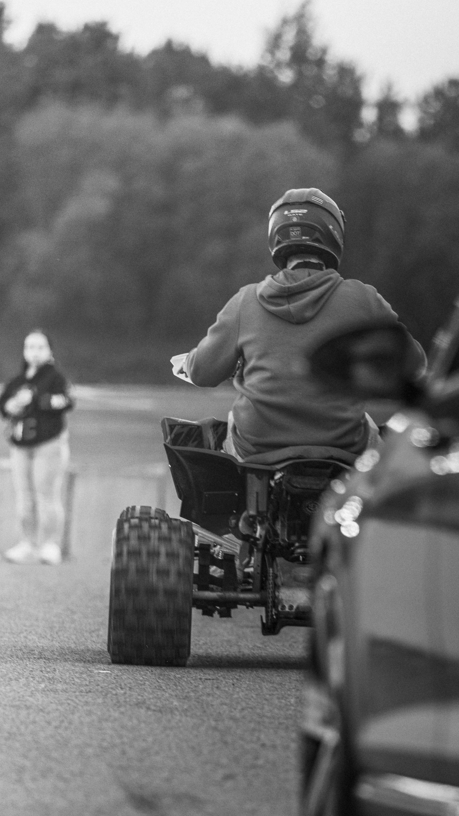Black and white rear view of a helmeted rider on a quad bike on a track while a spectator watches in the background.