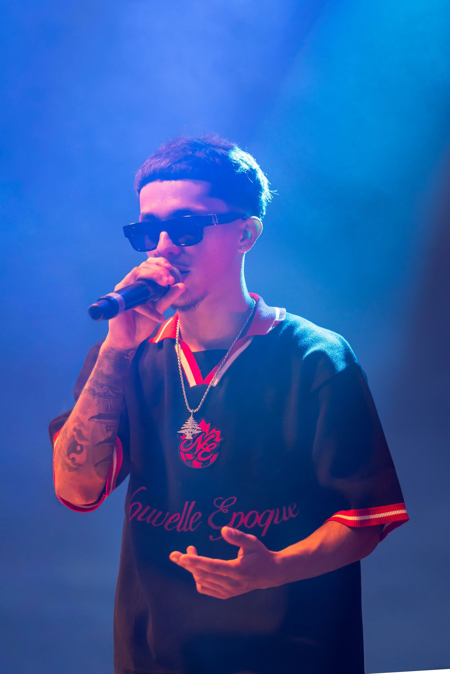Rapper in dark sunglasses and a black Nouvelle Epoque jersey performs into a microphone under blue stage lighting.