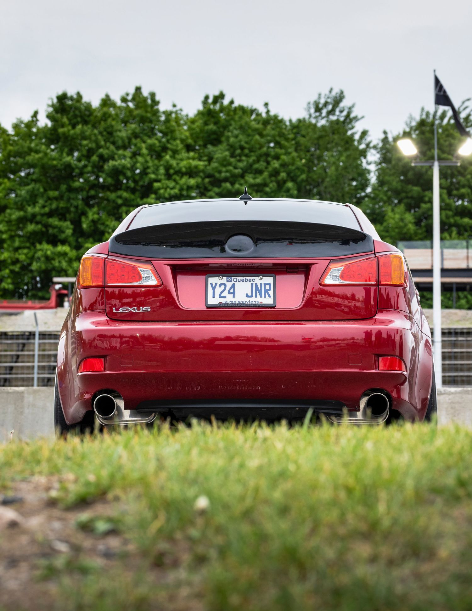 Rear view of a red Lexus sedan with dual exhaust tips and a Quebec license plate, parked on grass with green trees in the background.