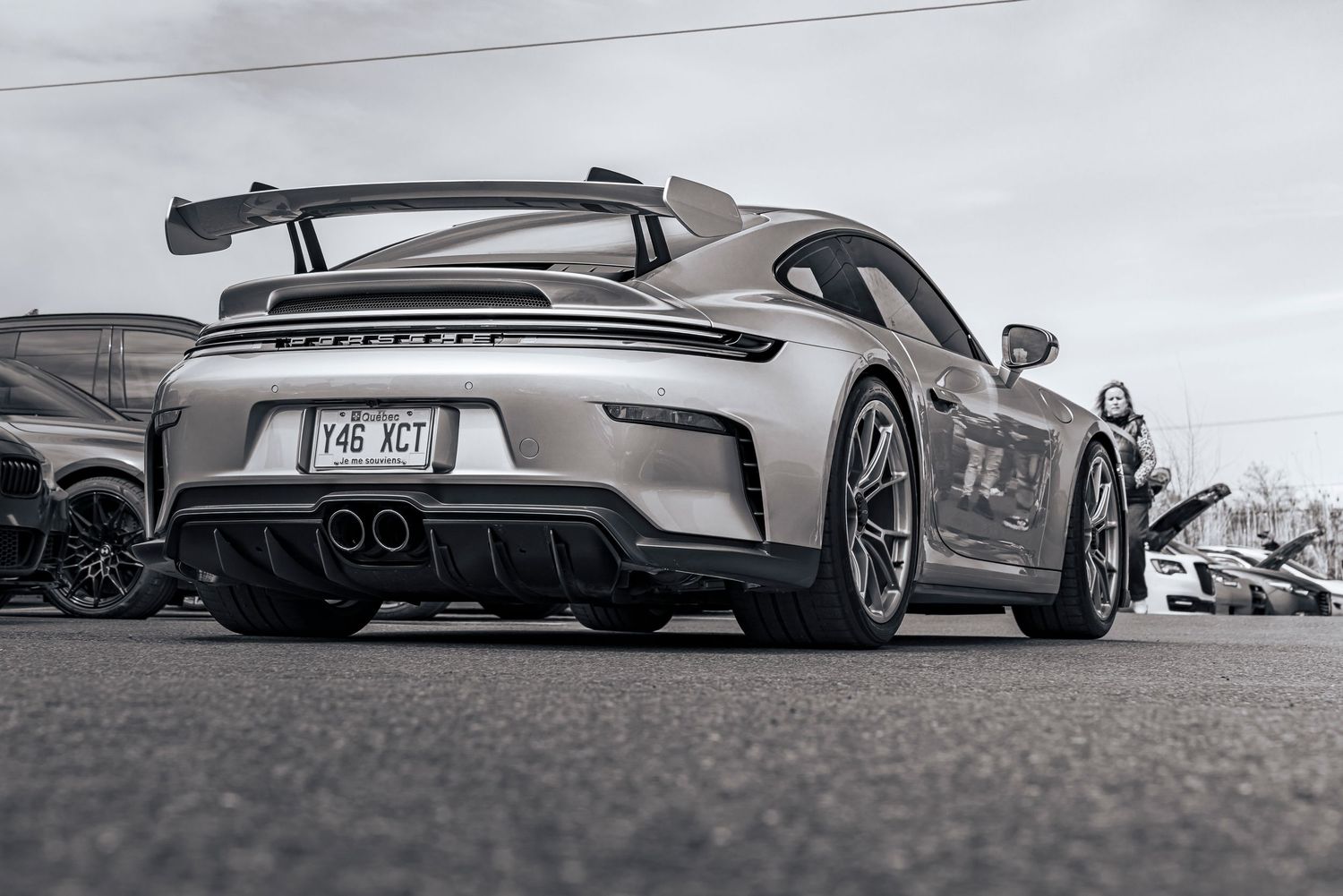 Rear view of a silver Porsche sports car with a large rear wing and dual exhaust parked in a lot at a car show.