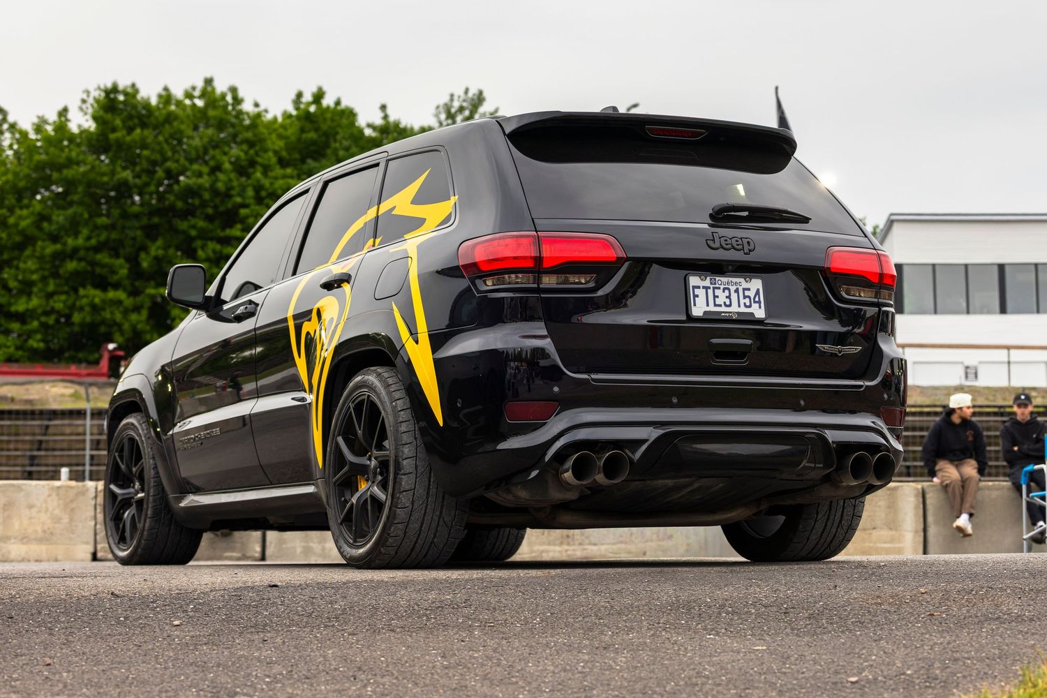 Black Jeep Grand Cherokee Trackhawk with yellow lightning bolt graphics and quad exhaust tips parked on a track with spectators behind.