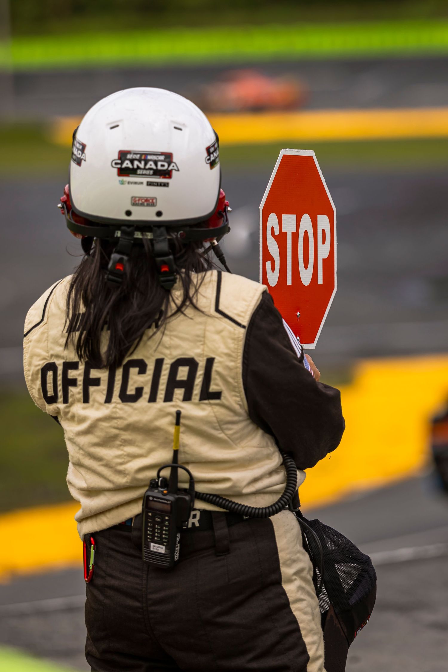 Race official in a white helmet and tan jacket holding a red stop sign trackside during a motorsport event.
