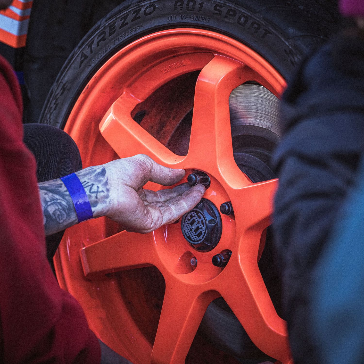 Tattooed hand tightens a lug nut on a bright orange racing wheel fitted with a sport tire.