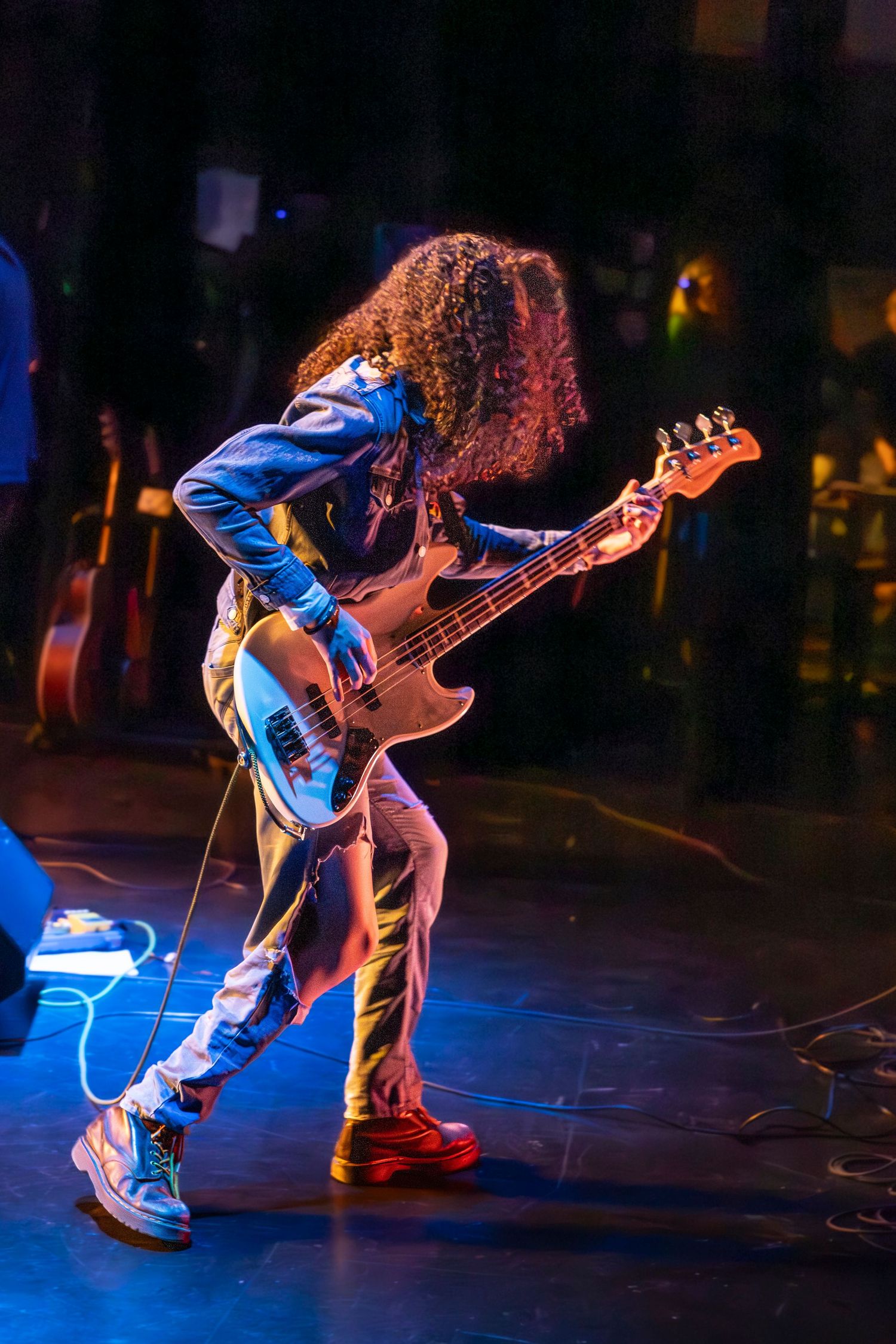 Bassist with long curly hair leans forward while playing an electric bass guitar on a stage lit in blue light.
