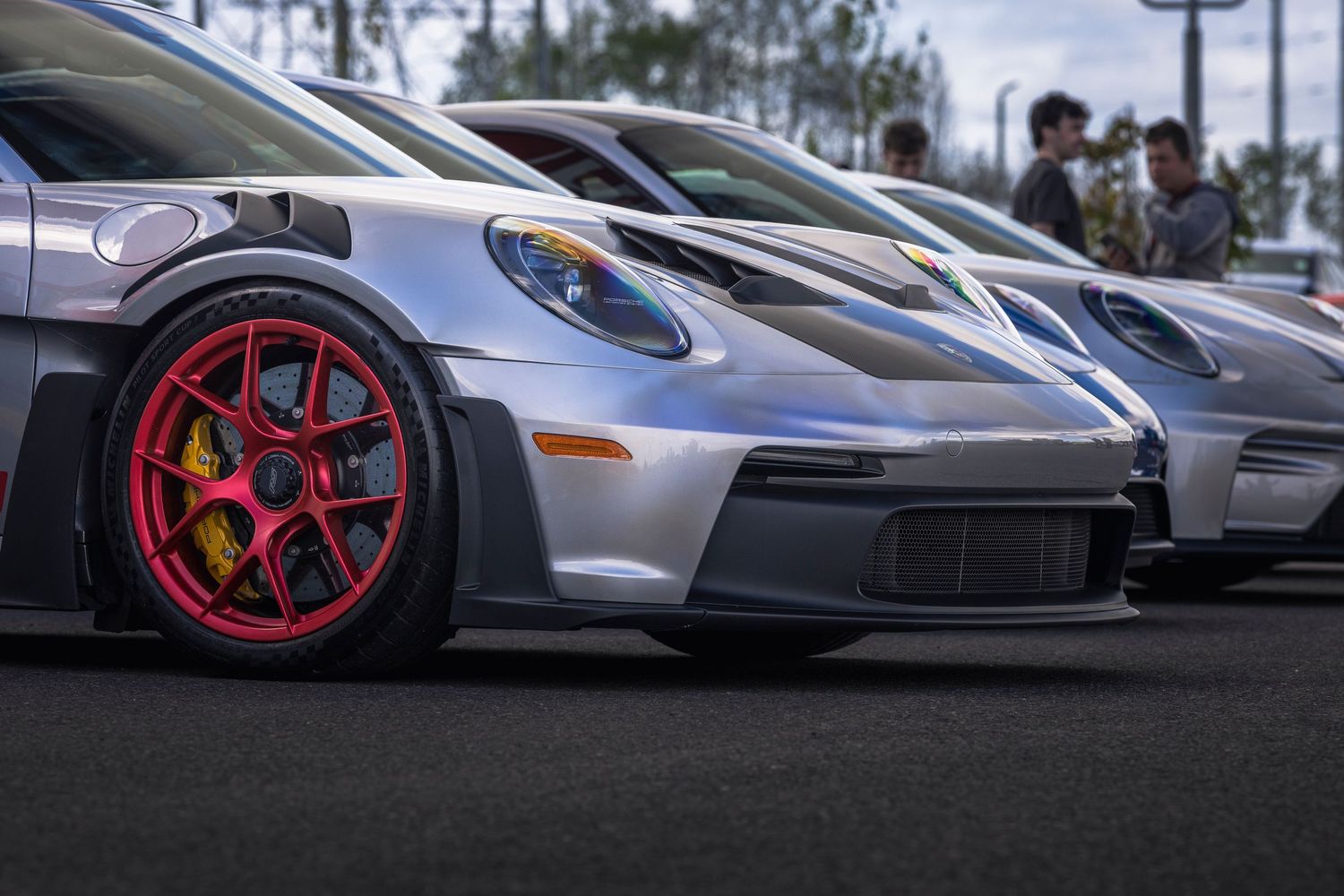 A row of silver Porsche sports cars with red and gold wheels parked at an outdoor car meet while spectators look on.