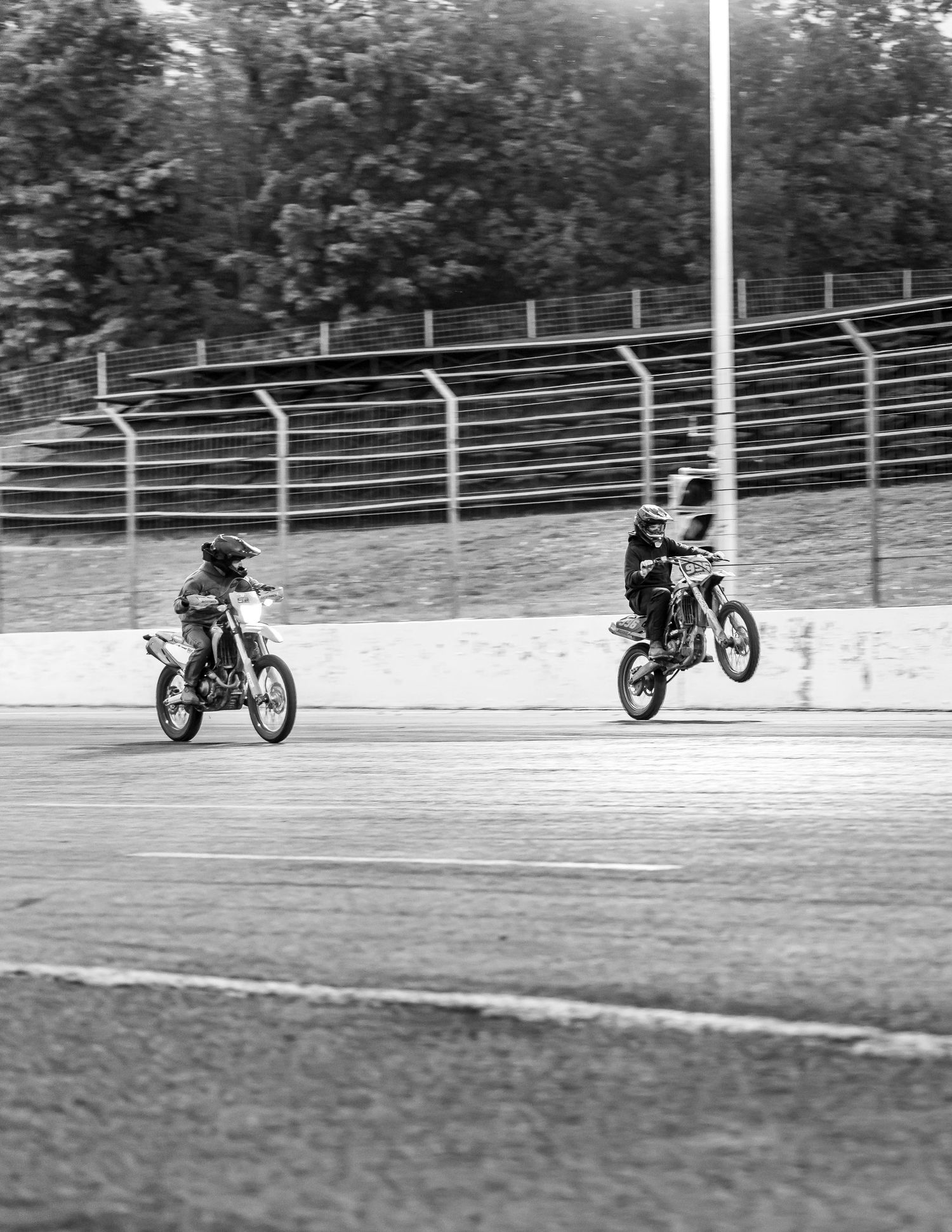 Black and white photo of two motocross riders popping wheelies on a race track in front of empty grandstands.