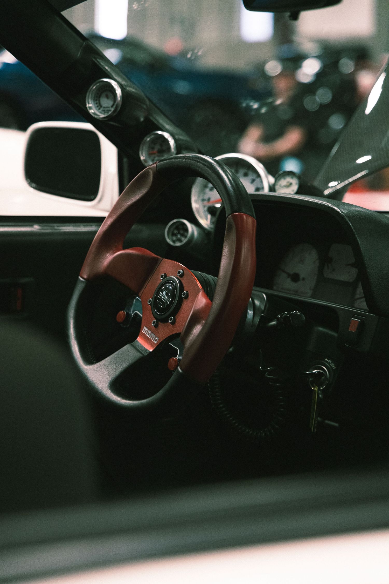 Interior view of a classic car showing a red and black sport steering wheel, auxiliary gauges and keys in the ignition.