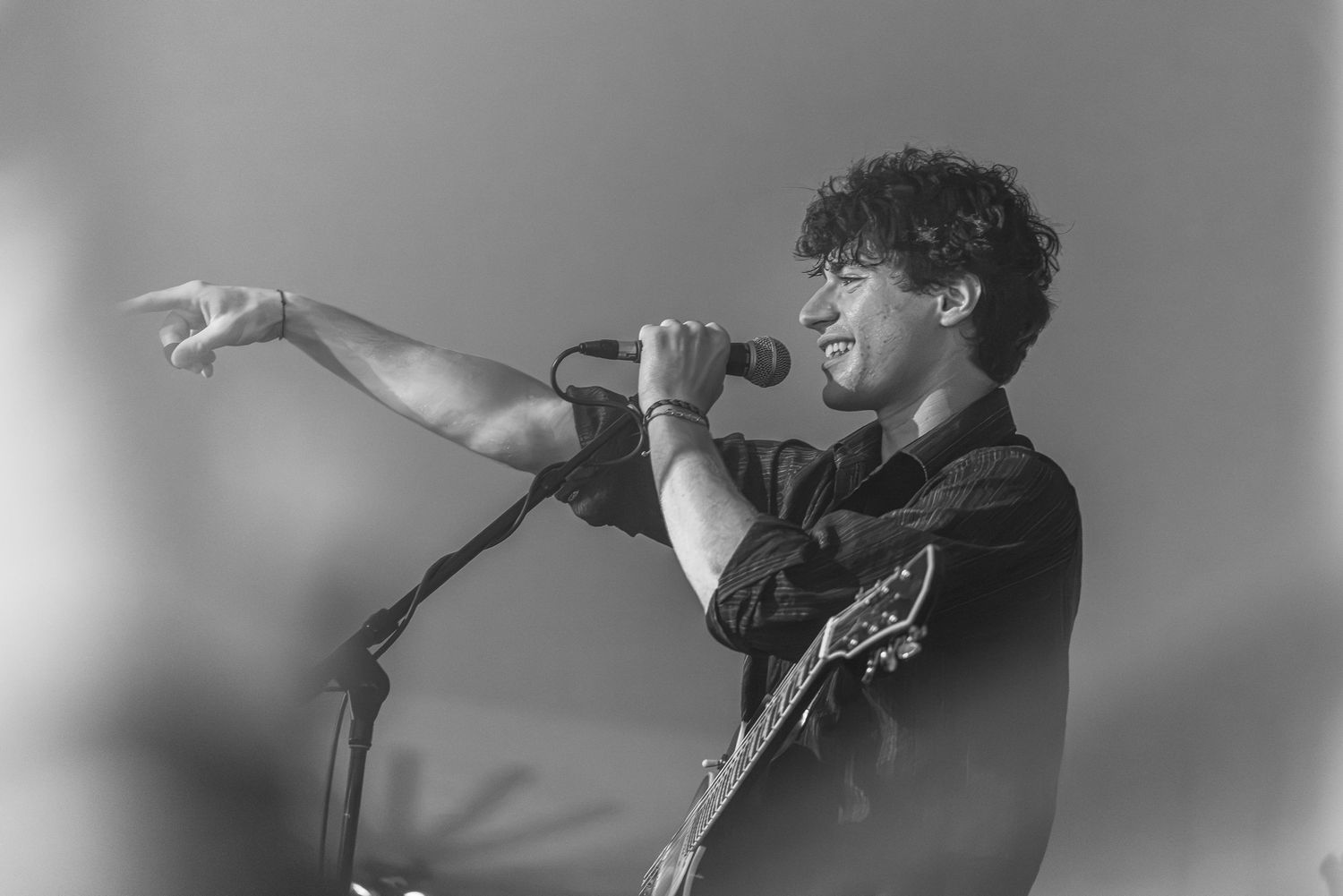 Black and white photo of a curly-haired guitarist singing into a microphone while pointing out toward the crowd during a live concert.