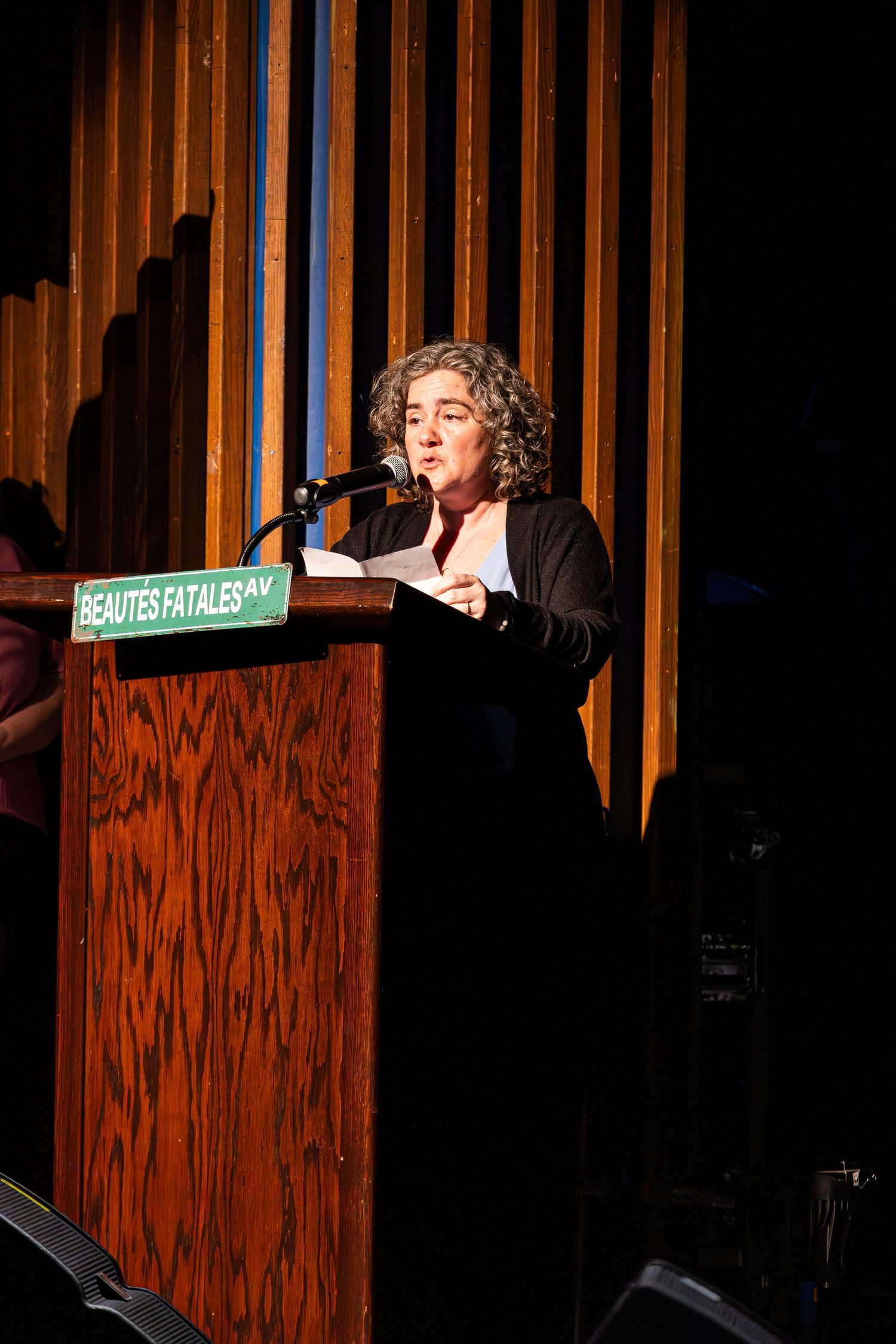 A woman with curly hair reads into a microphone at a wooden podium marked Beautés Fatales on a stage.