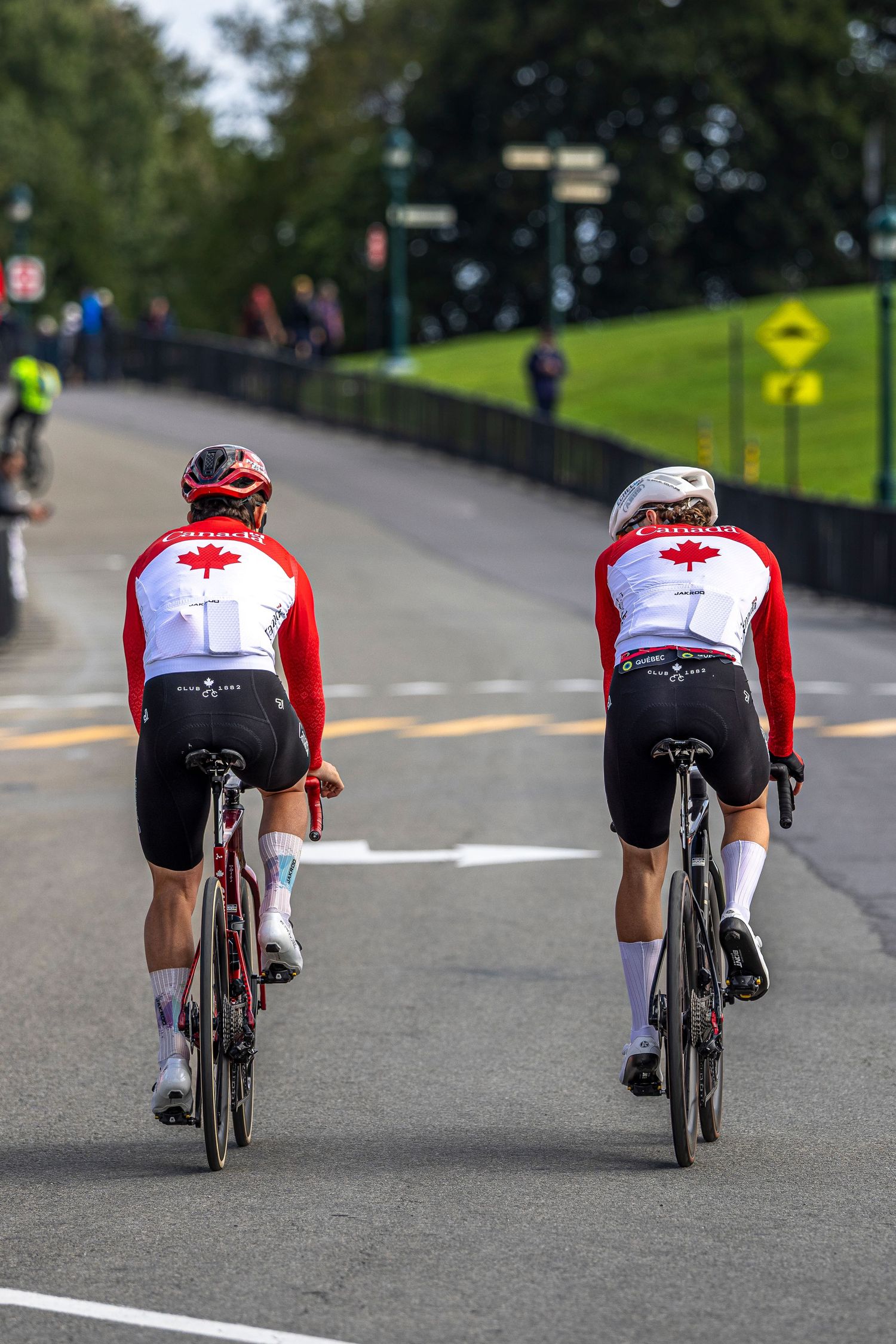 Two road cyclists in red and white Canada jerseys and helmets ride bicycles along a paved road near a grassy hill.