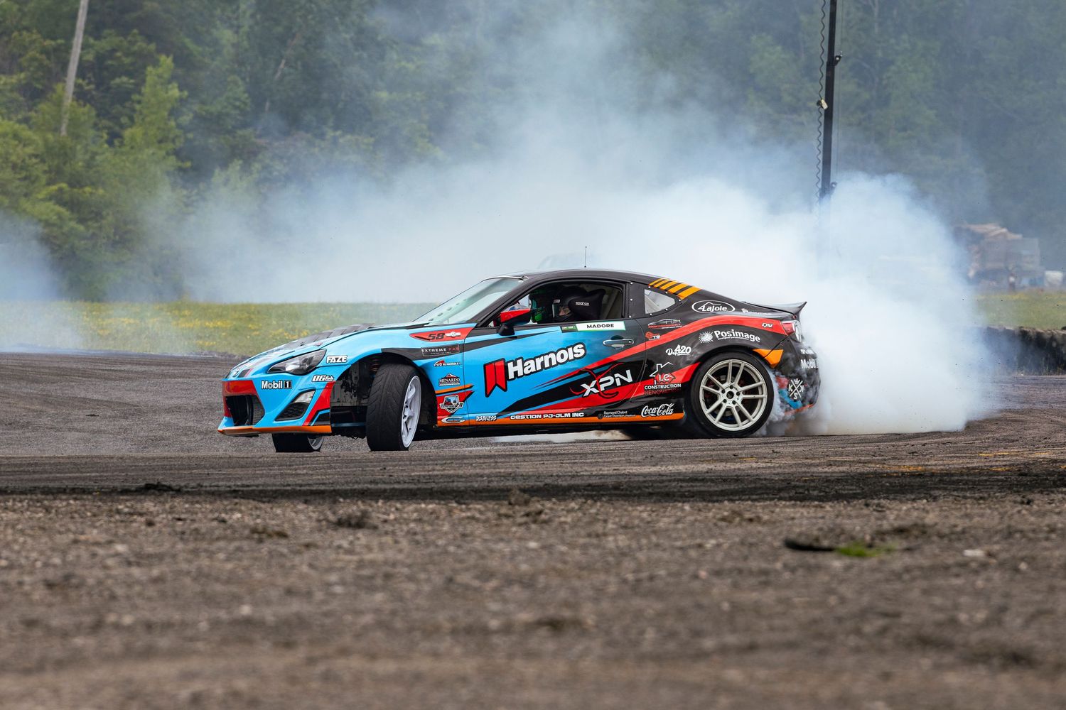 Blue and red sponsor-covered drift car sliding through a corner on a dirt-edged track as white smoke rises behind it.