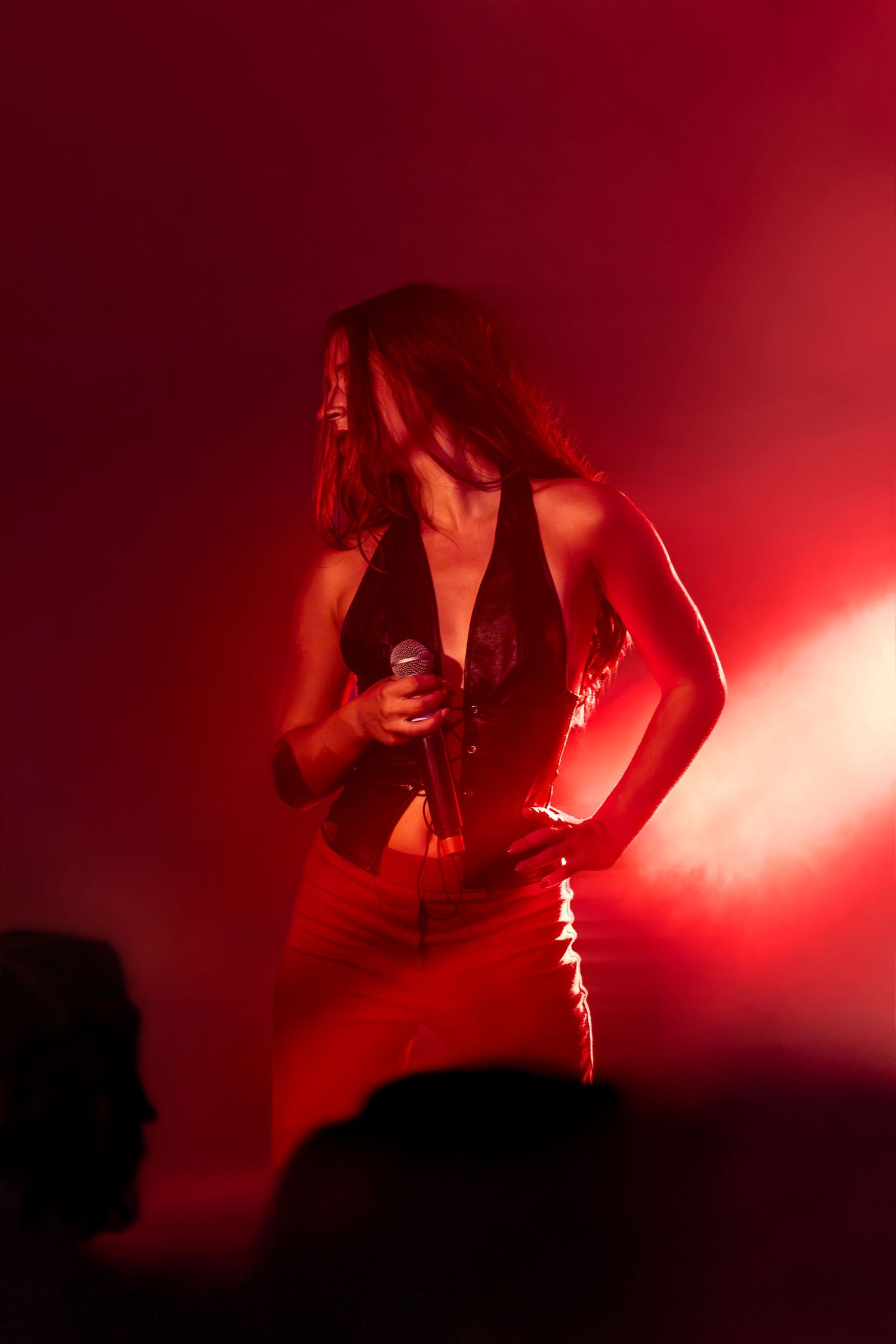 Silhouetted female vocalist in a black top and red pants holds a microphone on stage, bathed in dramatic red stage lighting and haze.