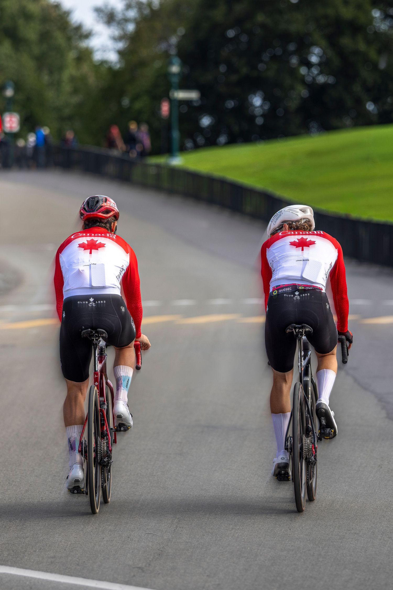 Two cyclists in red and white Canada jerseys ride road bikes along a paved curving road beside a grassy hill.
