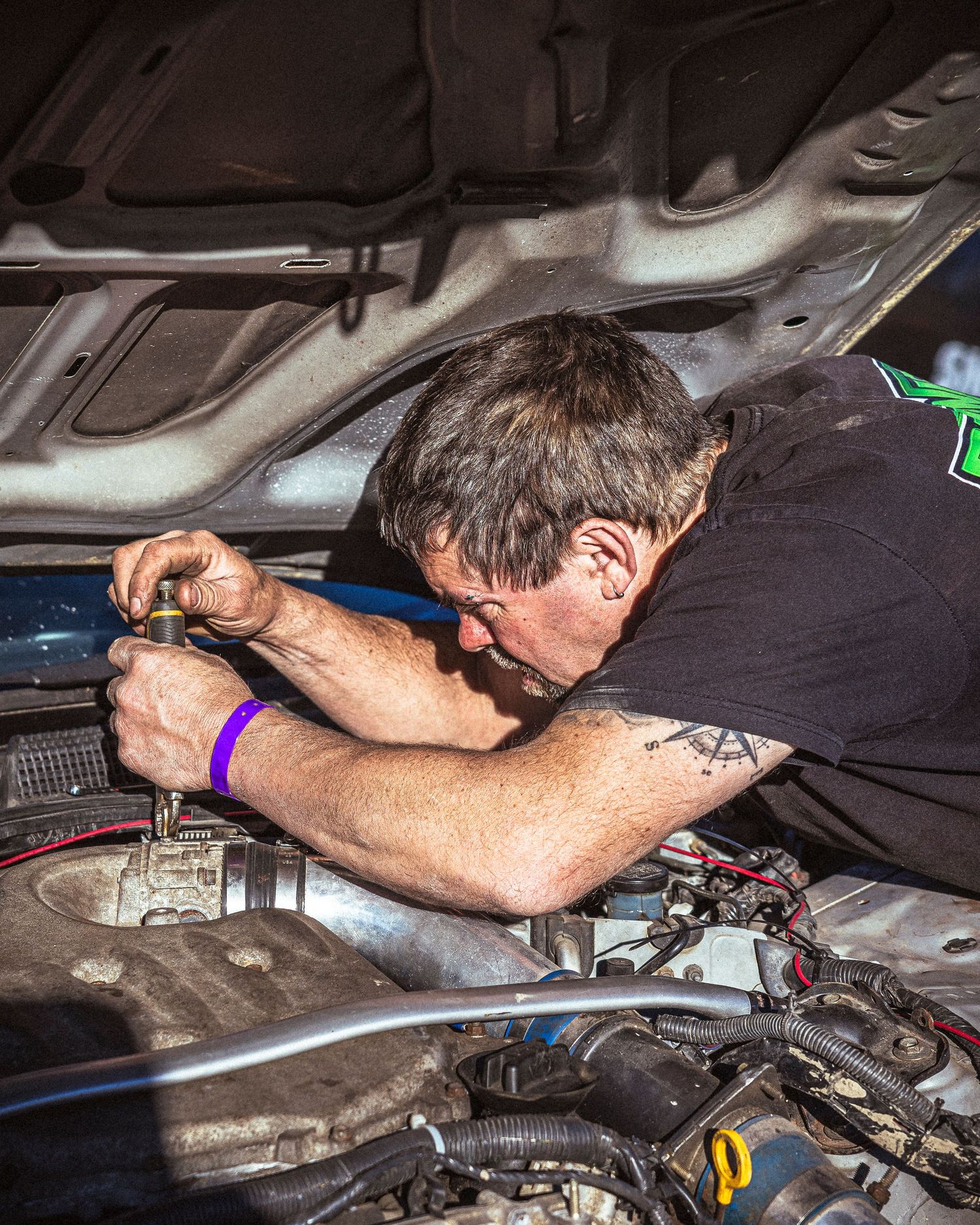 Mechanic in a black t-shirt leans over an open hood and works on a car engine with a small tool.
