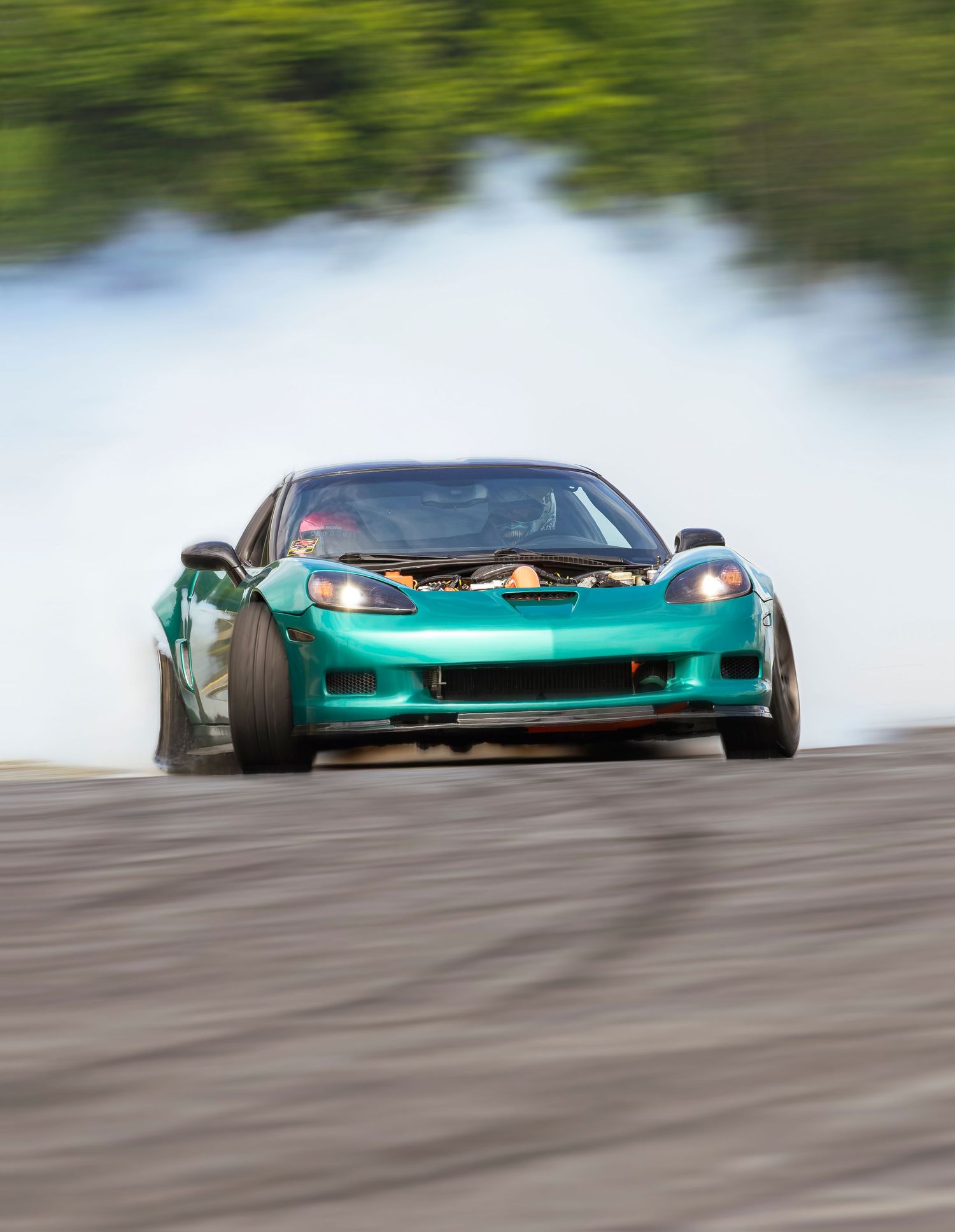 Teal green sports car sliding sideways in a drift with white tire smoke and blurred greenery behind it.