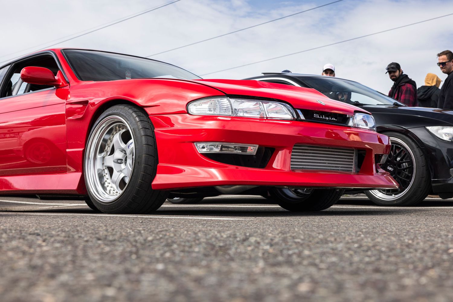 Low-angle view of a red Nissan Silvia S14 with aftermarket silver wheels and a front-mount intercooler at a car meet with spectators behind.