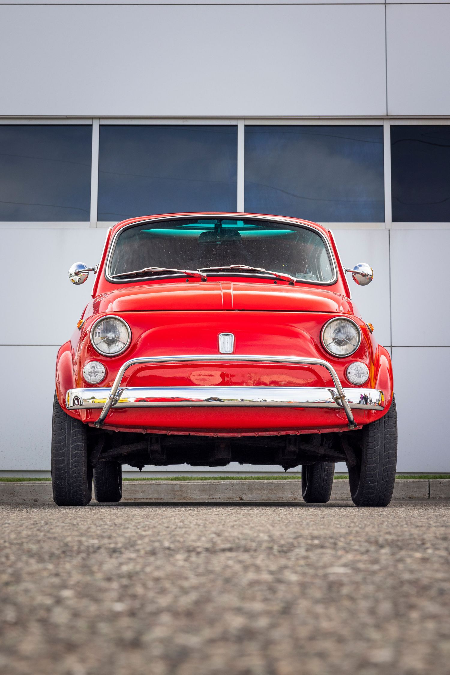 Front view of a vintage red Fiat 500 with chrome bumper and round headlights parked on gravel in front of a modern building.
