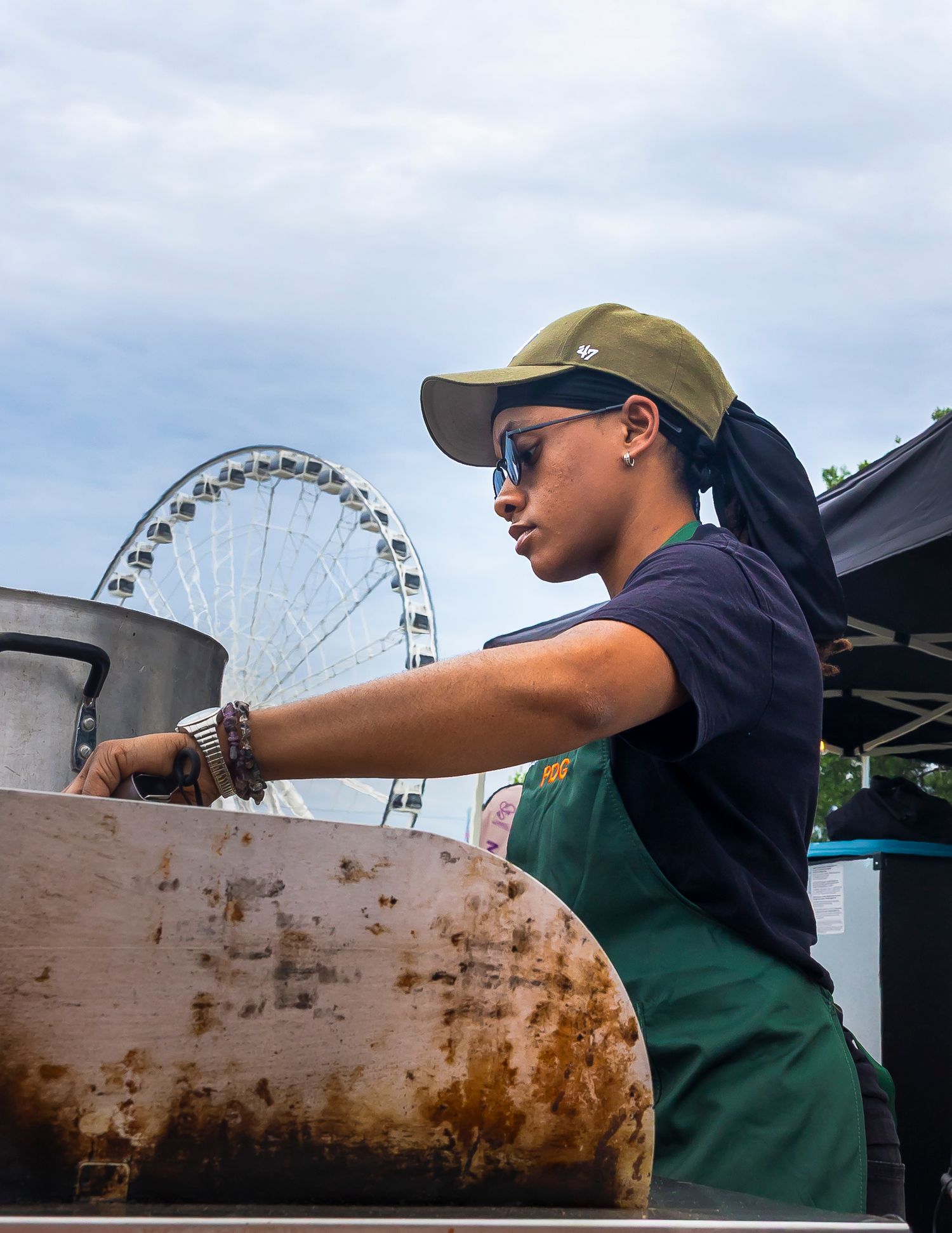 A woman in a green apron and cap works at a food stand with a large Ferris wheel visible in the background.