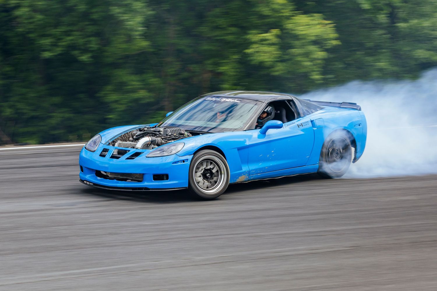Bright blue sports car with an open hood drifting on a wet track as smoke pours from the rear tire.