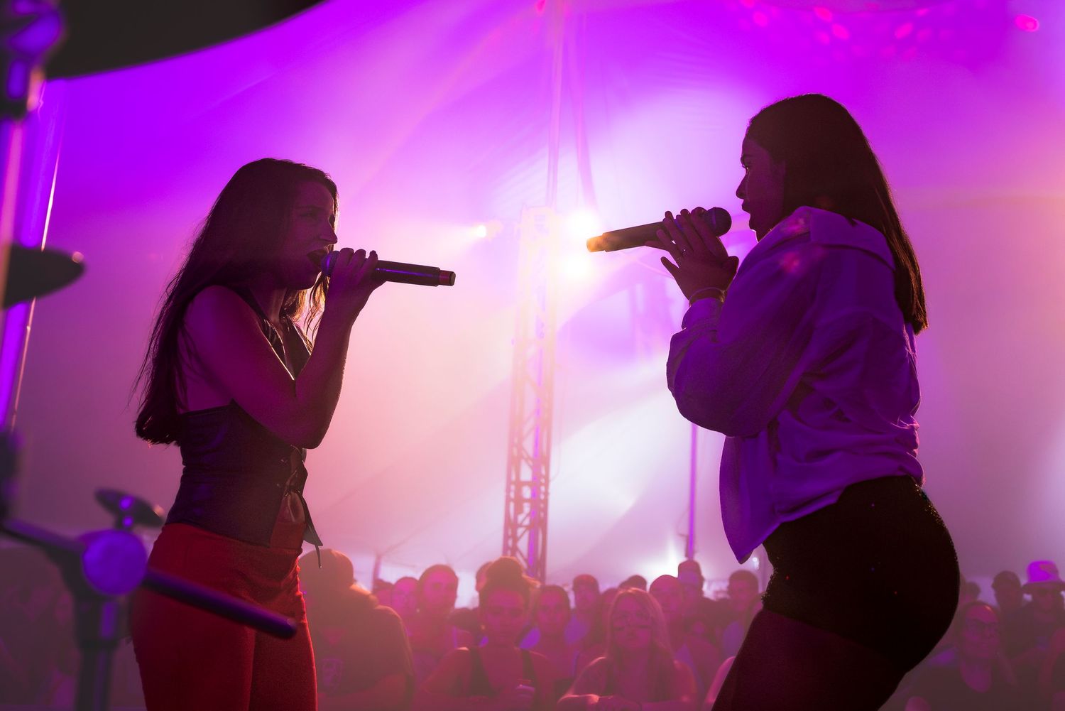 Two women sing into microphones on stage under bright magenta lights, with a crowd silhouetted in the background at a live music event.