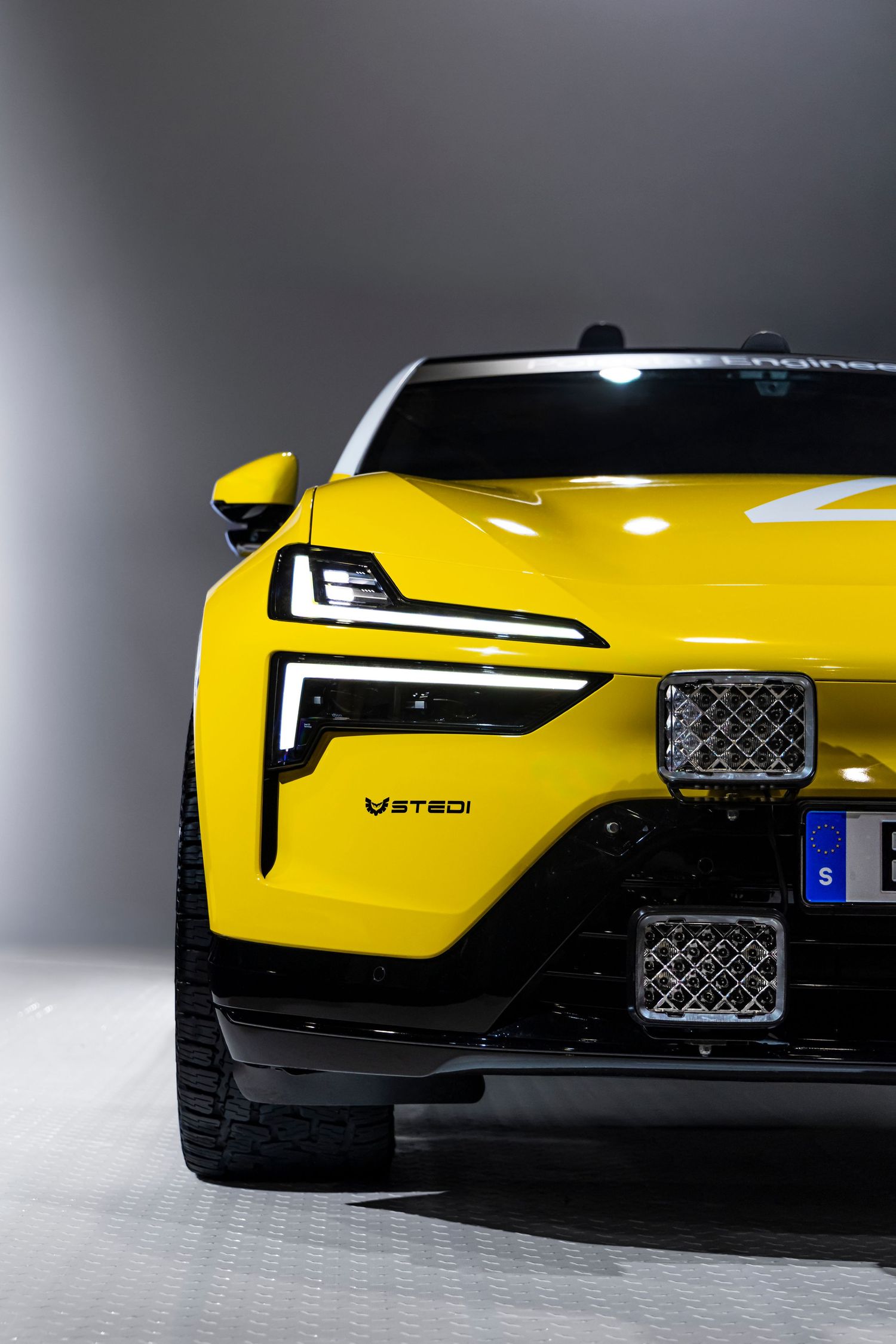 Front close-up of a bright yellow electric SUV with sleek LED headlights and mesh grille inserts in a studio setting.