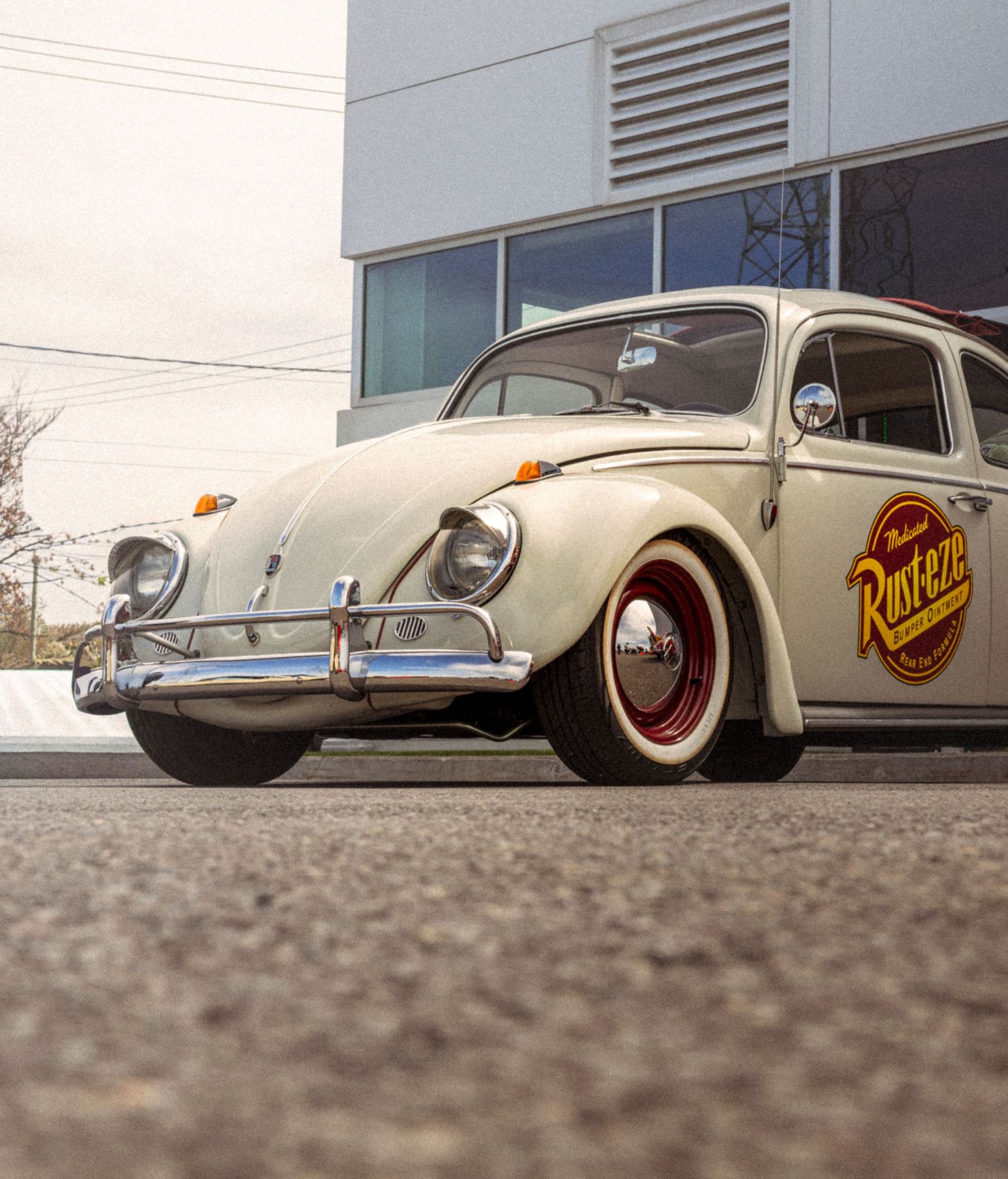 Cream vintage Volkswagen Beetle with chrome bumper, red wheels and a round Rust-Eze logo parked on asphalt in front of a modern building.
