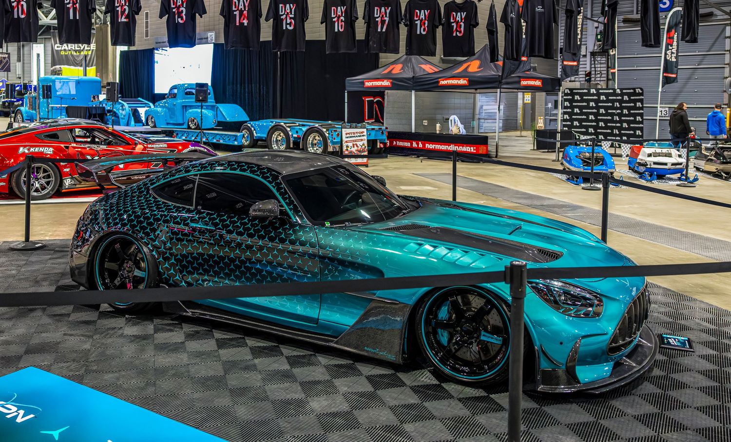 A teal Mercedes-AMG GT sports car with a sculpted body is displayed on a roped-off stand at an indoor automotive show.