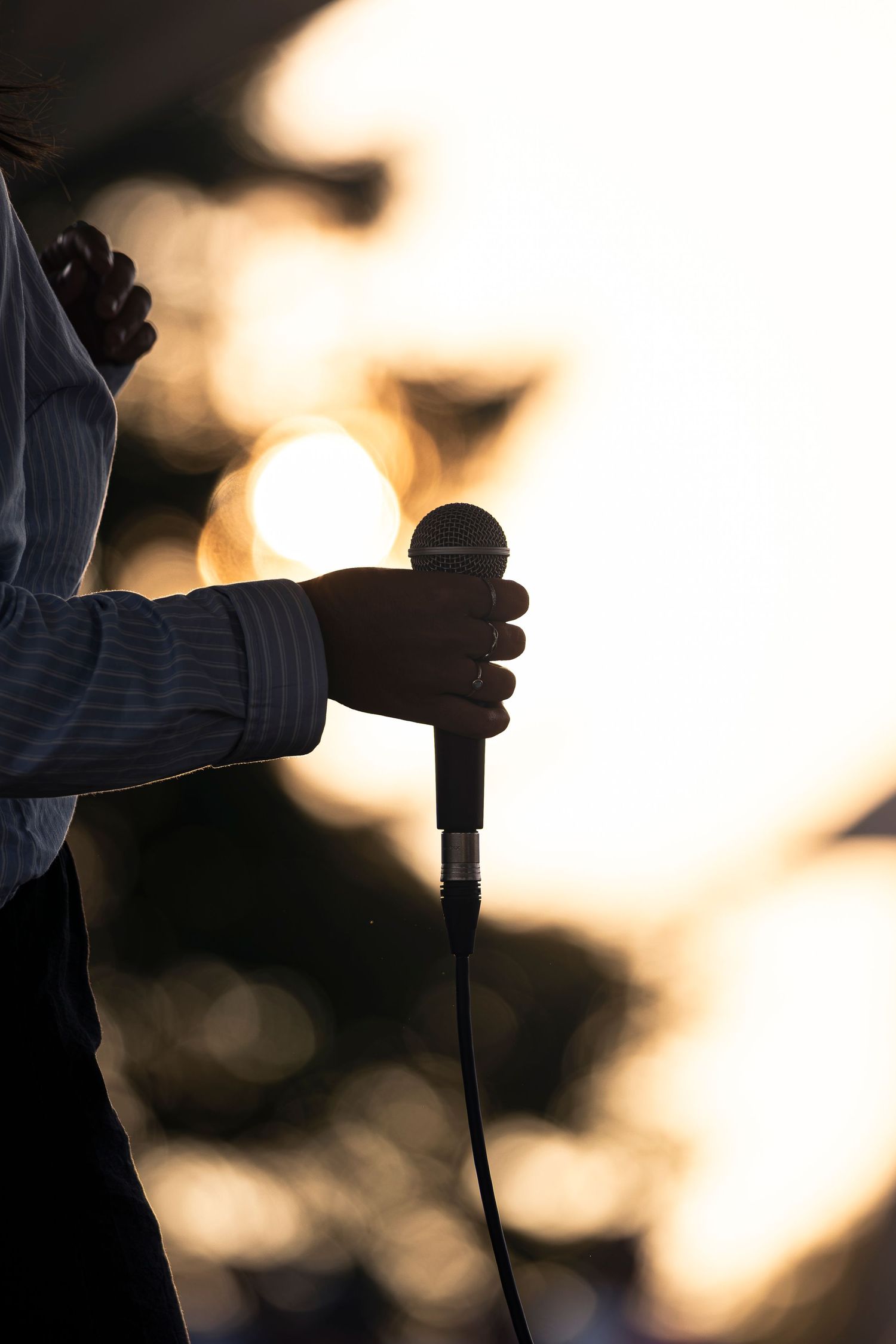 Silhouette of a hand gripping a microphone with cable against a glowing, softly blurred golden sunset background.