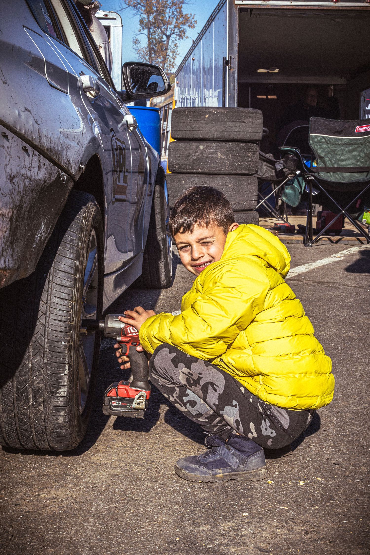Smiling boy in a yellow puffer jacket crouches beside a car and uses a cordless impact wrench on a wheel.