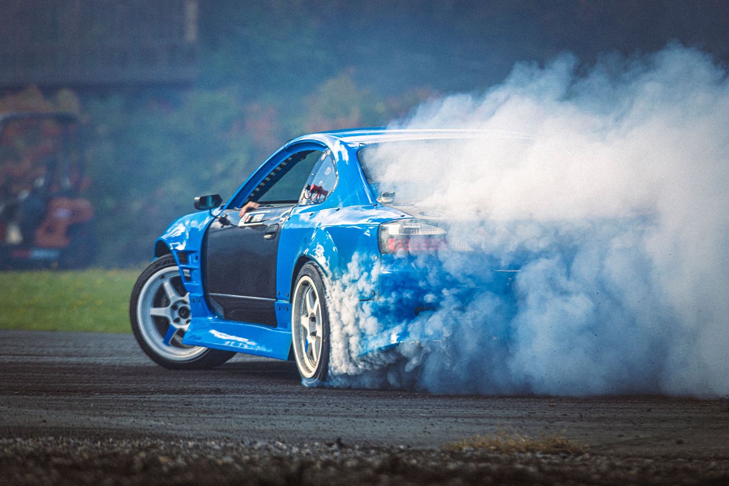 Blue drift car sliding sideways on asphalt as thick white tire smoke billows behind it.