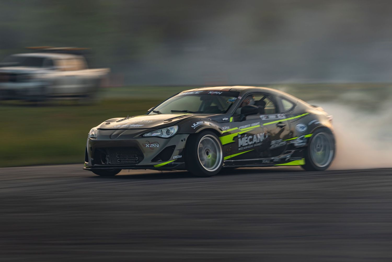 Black and green sponsored drift car sliding through a turn on a racetrack with light tire smoke behind it.