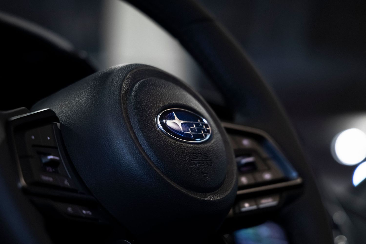Close-up of a Subaru steering wheel with the blue star logo on the center airbag cover and control buttons in a dark car interior.