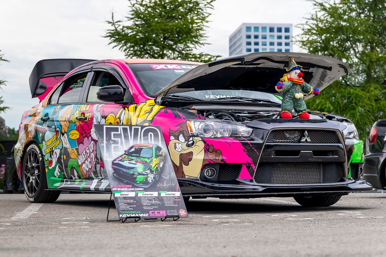 Colorful cartoon-wrapped Mitsubishi Lancer Evolution with open hood and a small elf toy on display at an outdoor car show.