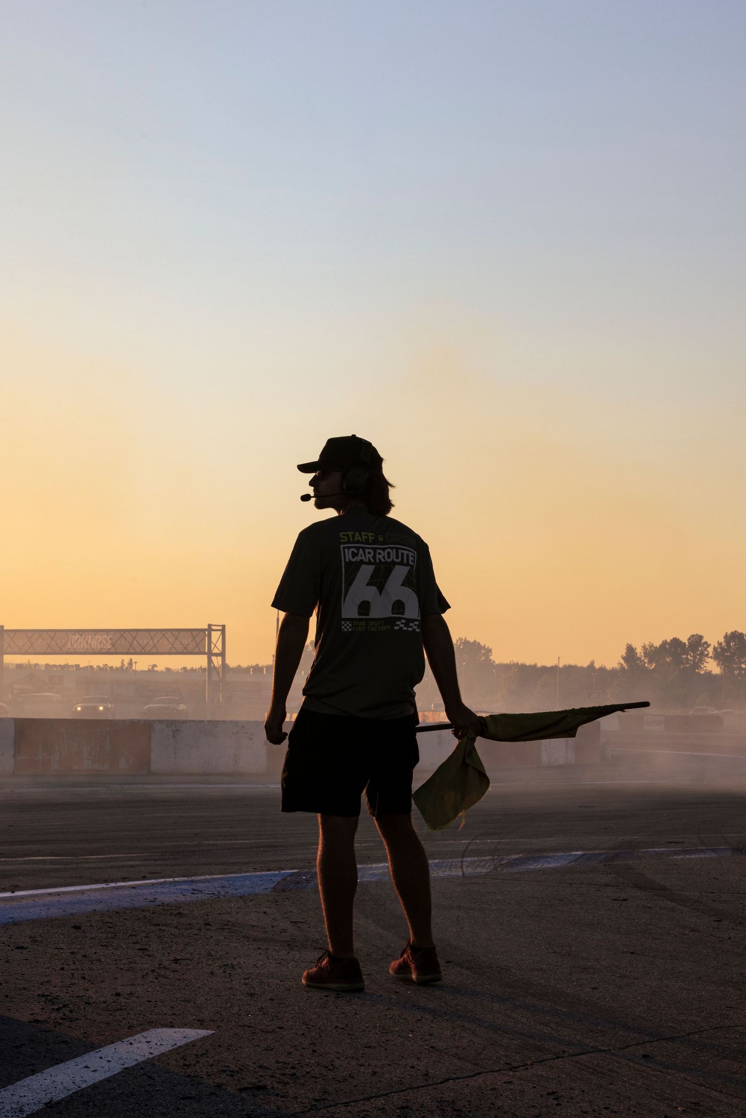 A track marshal wearing a headset and holding a flag is silhouetted against a glowing orange sunset sky at a racing event.