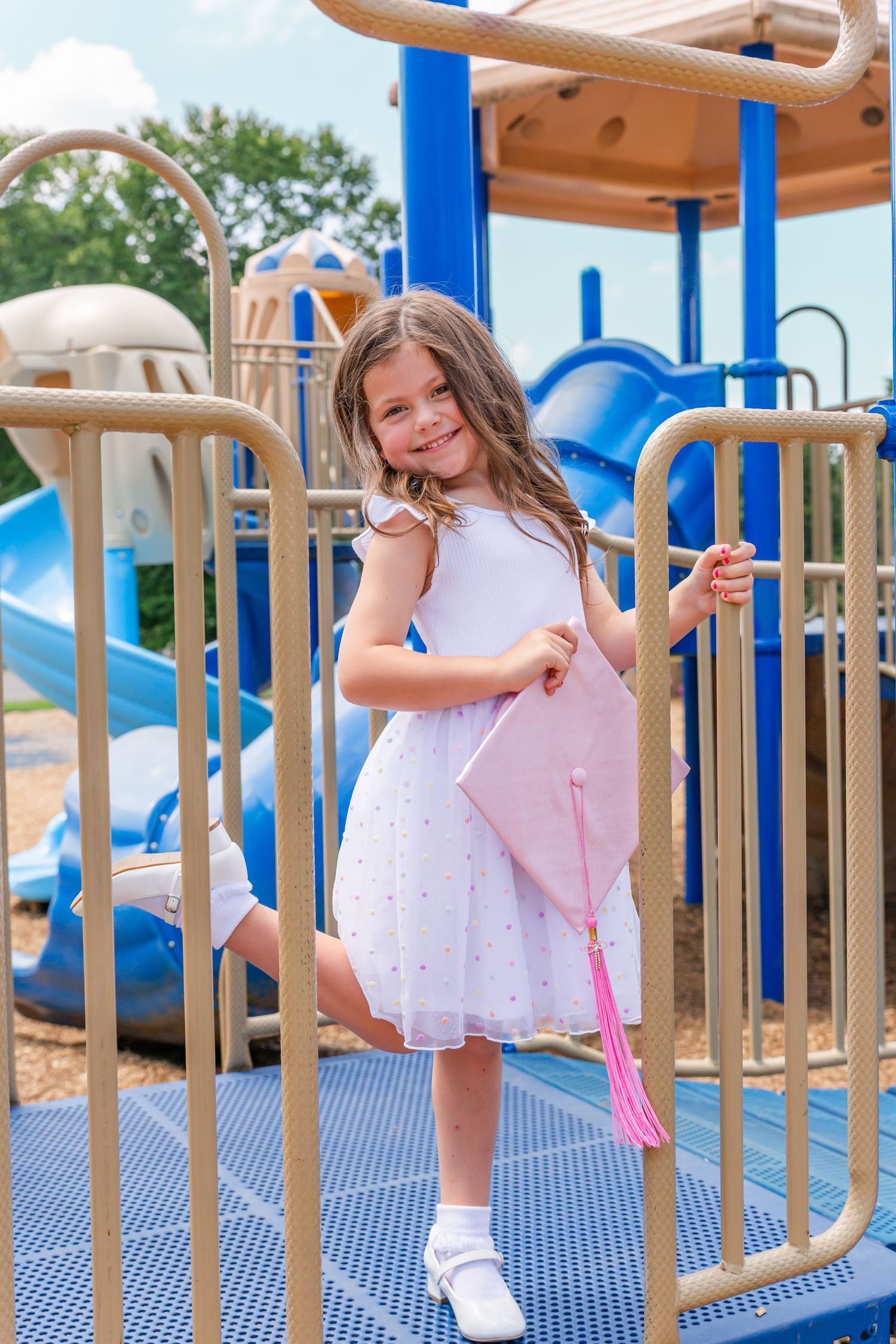 White dress and pink graduation gown twirl playfully on blue playground equipment.