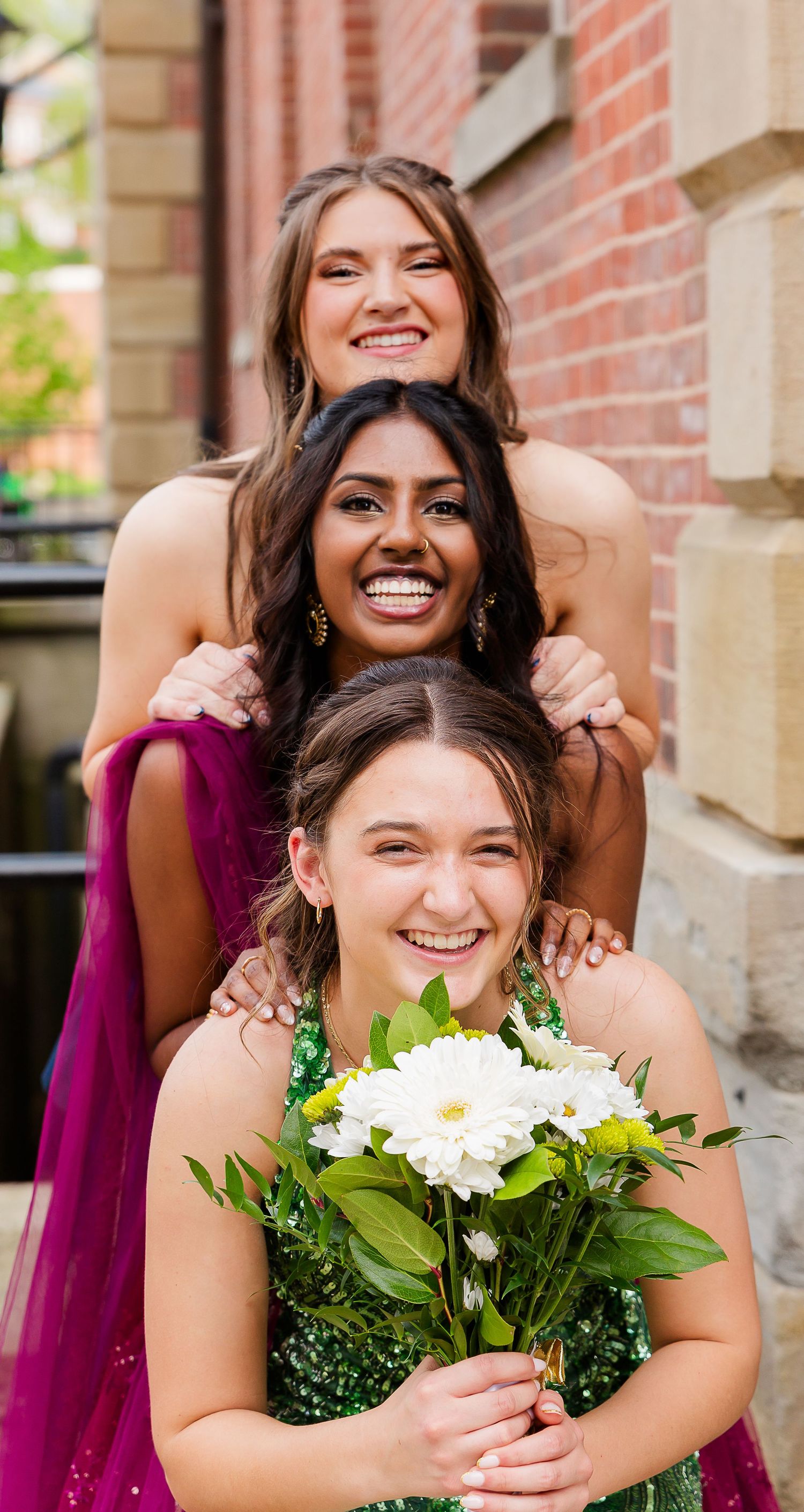 Three smiling young women in formal dresses pose together, one holding white flowers, near a brick wall.