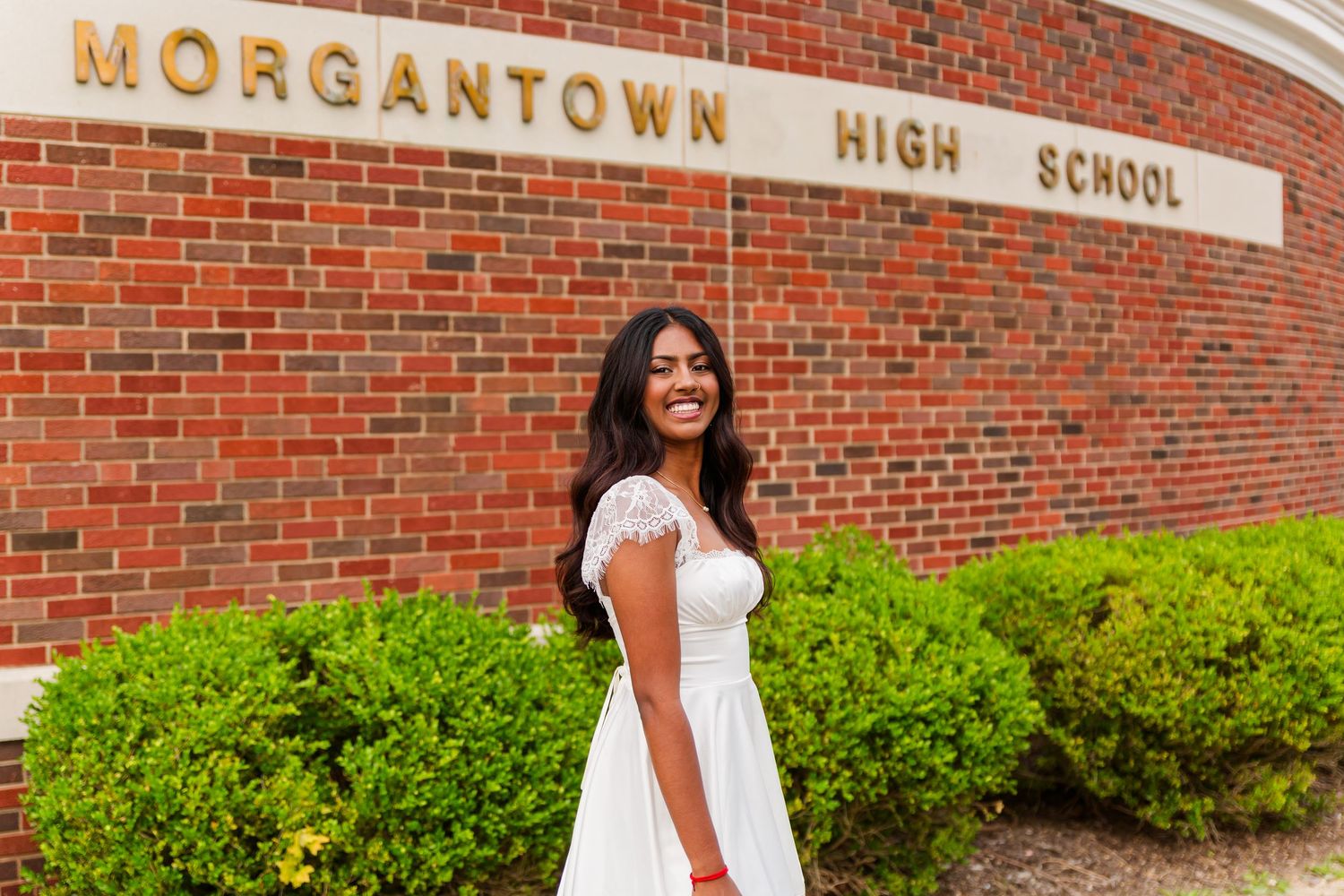 Smiling young woman in white dress posing in front of Morgantown High School brick wall with green shrubs.