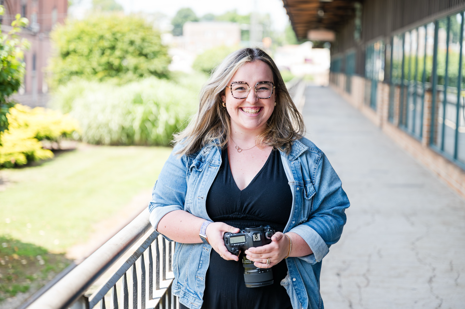 Smiling woman with glasses holding a camera outdoors, wearing a denim jacket over a black dress on a sunny day.