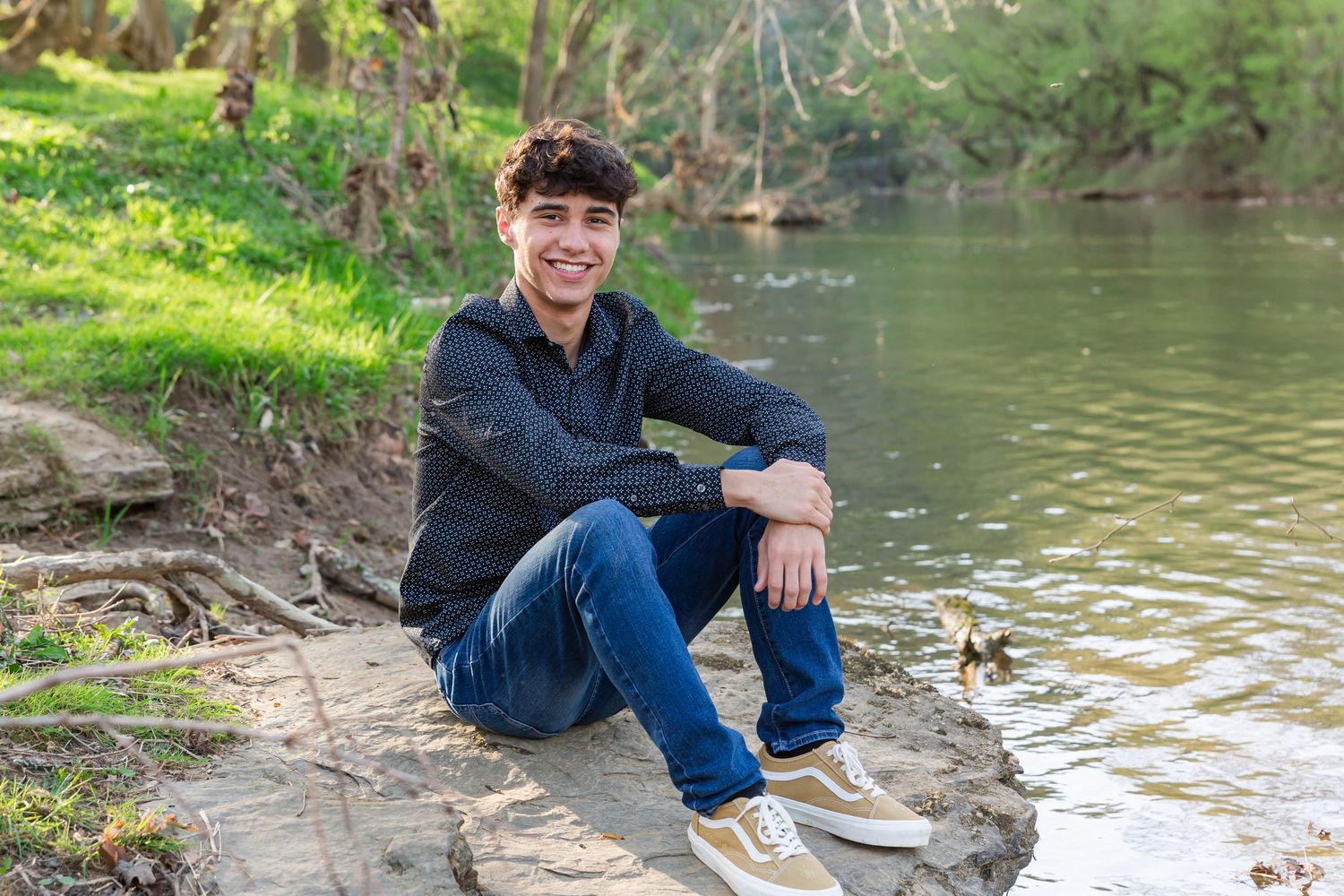 Student in dark shirt relaxes by riverside in casual outdoor portrait.