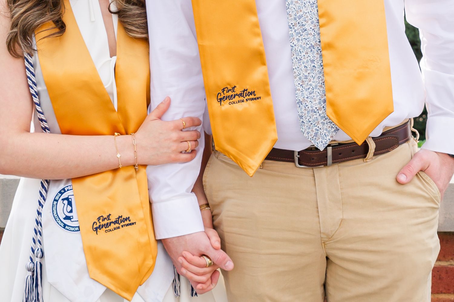 Row of graduates wearing gold stoles and khaki pants stand together during commencement.