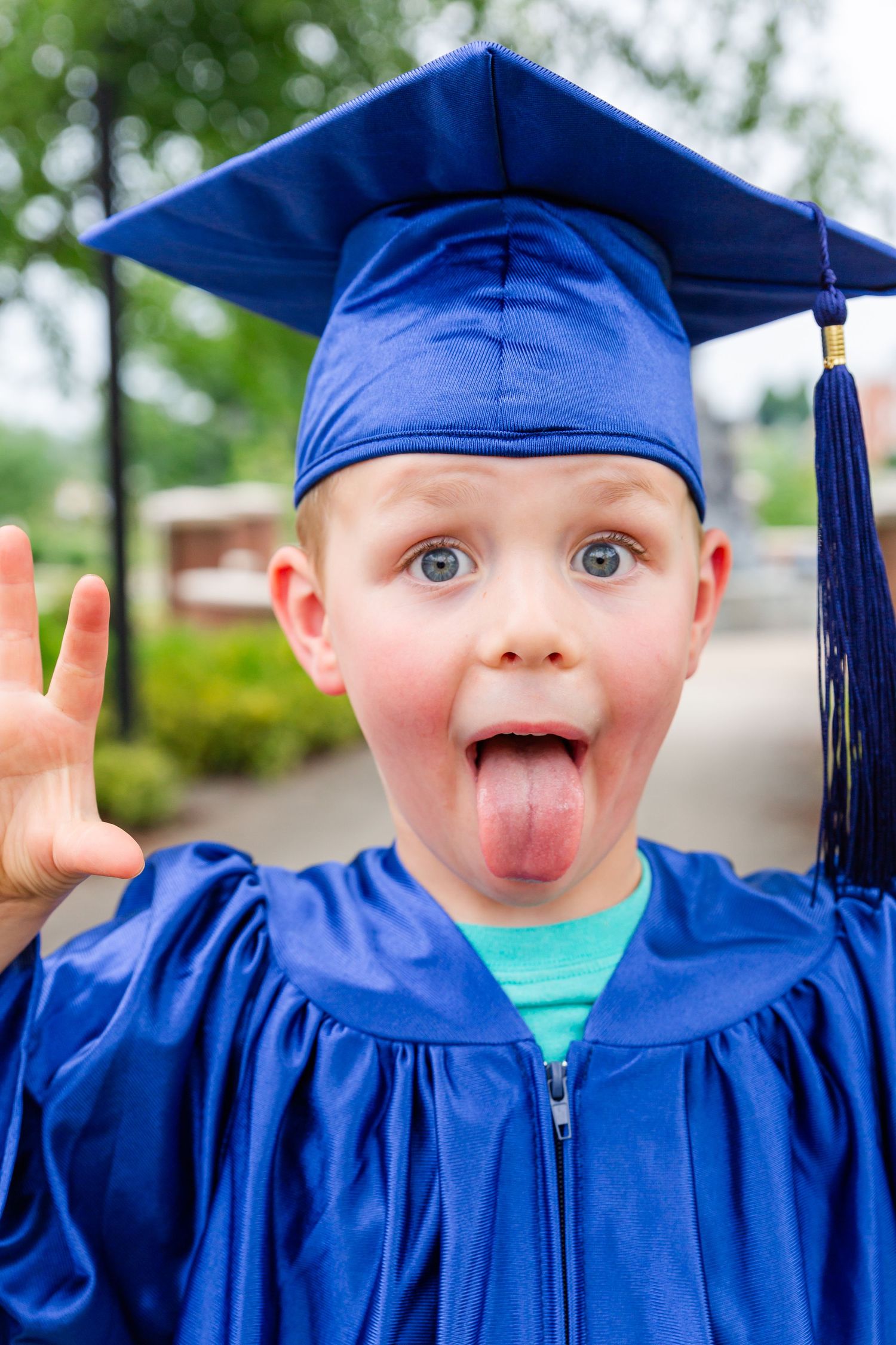 Young graduate in blue cap and gown makes silly expressions outdoors.
