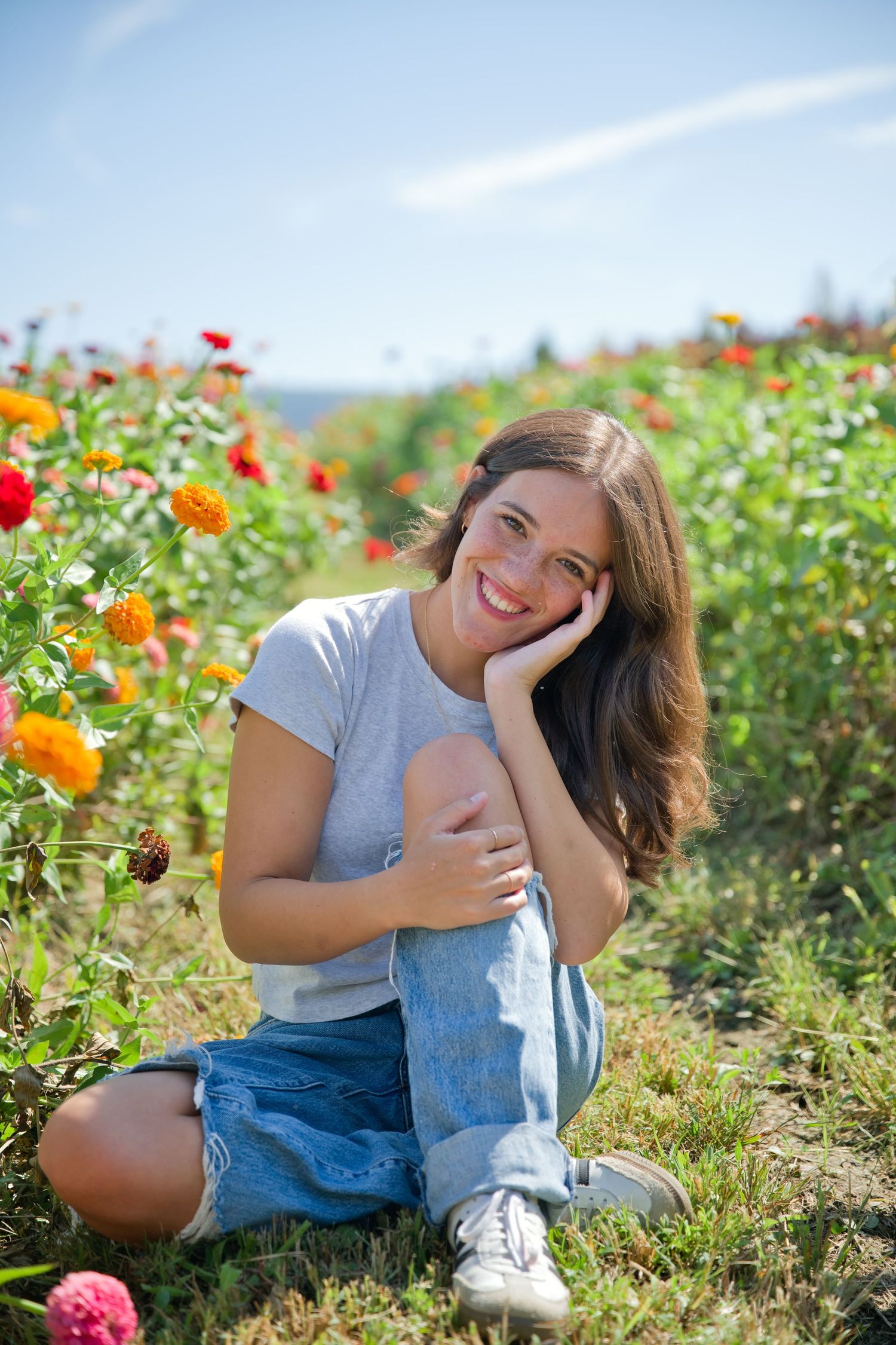 Young woman sitting in a colorful flower field, smiling cheerfully on a sunny day, wearing jeans and a gray t-shirt.