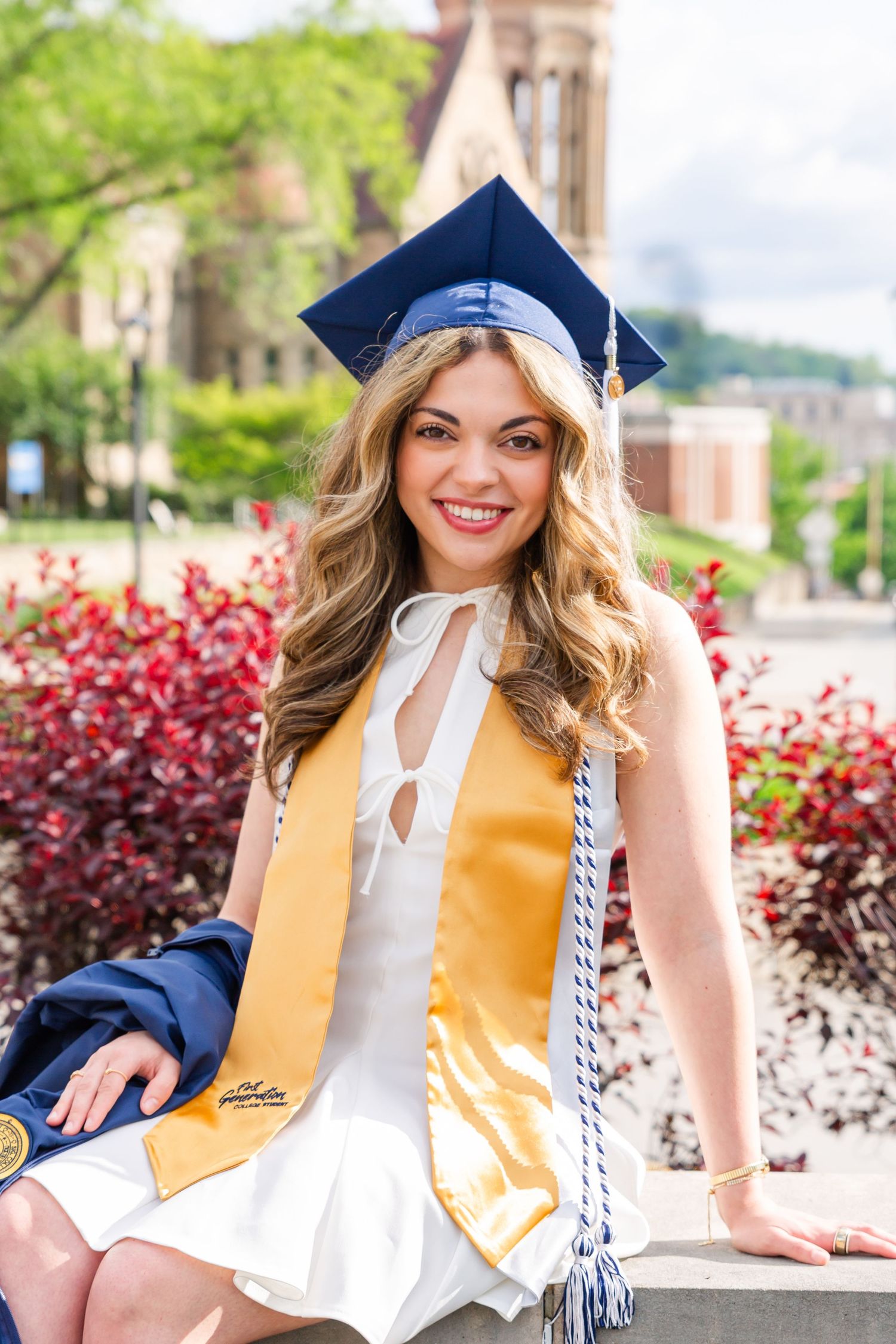 Graduate in white dress and gold stole poses outdoors with decorative red plants in background.