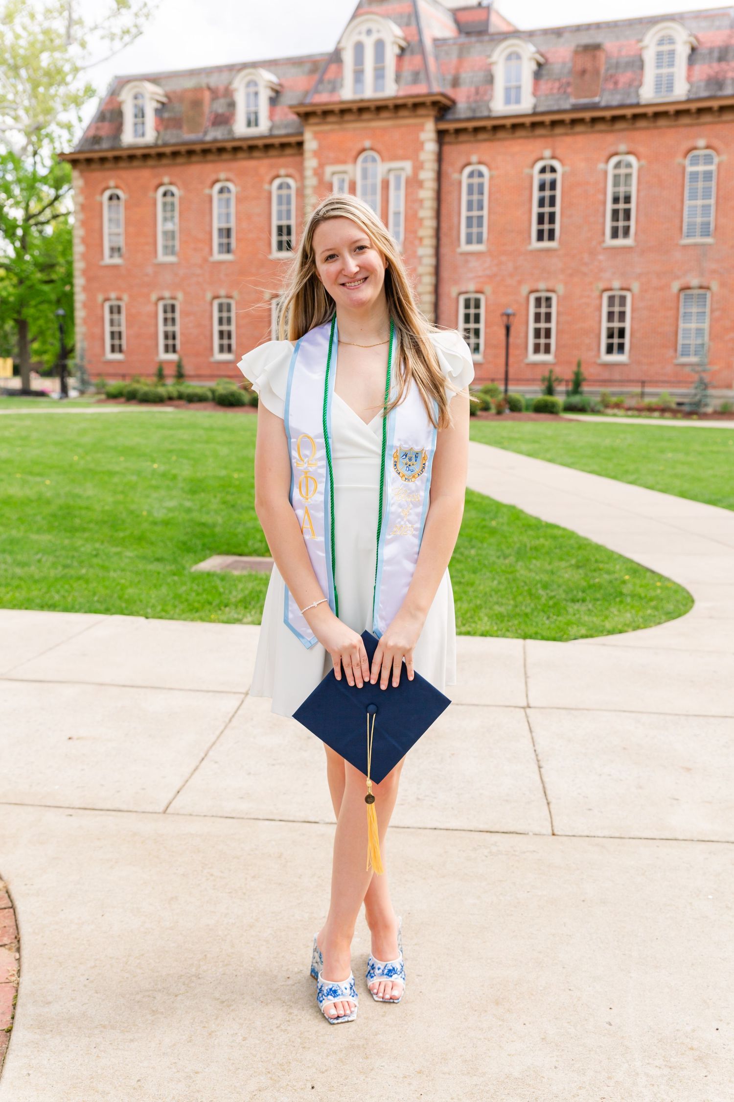 Graduate poses in front of historic brick building wearing light blue sash and navy shorts.