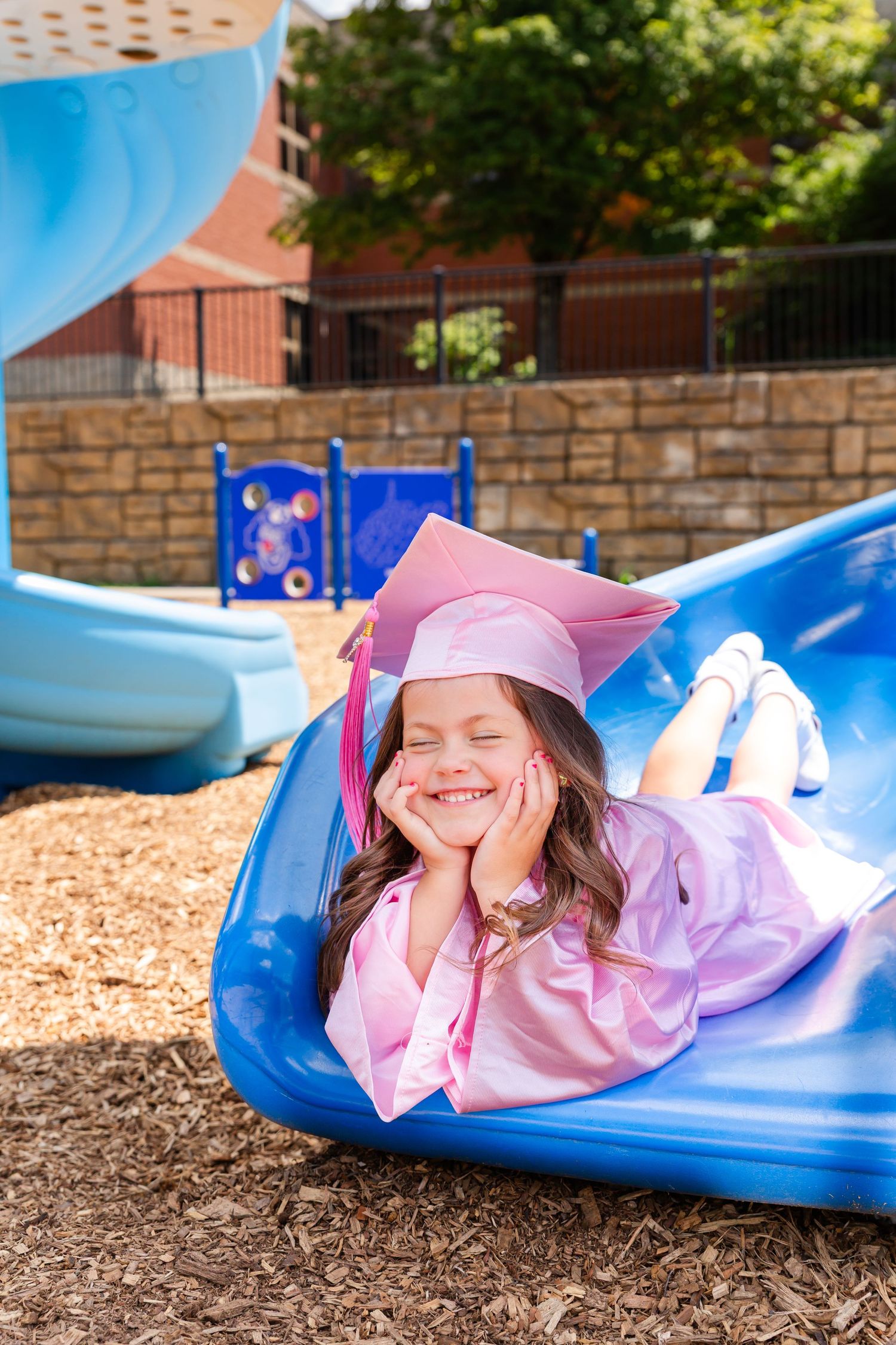 Graduate in pink dress laying playfully on a blue playground slide outdoors.