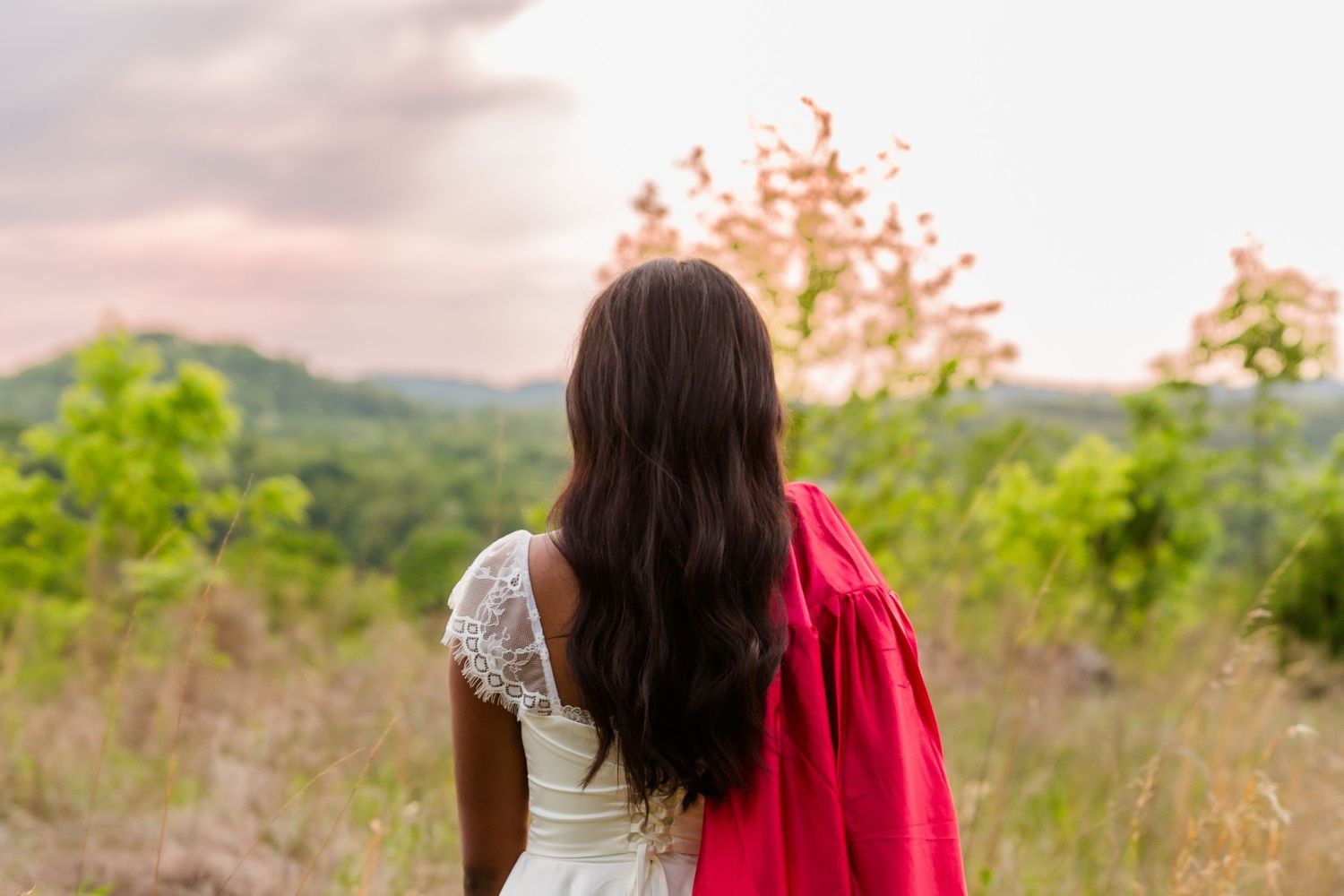 Young woman with long dark hair in white dress holding red fabric, gazing at lush green hills at sunset.